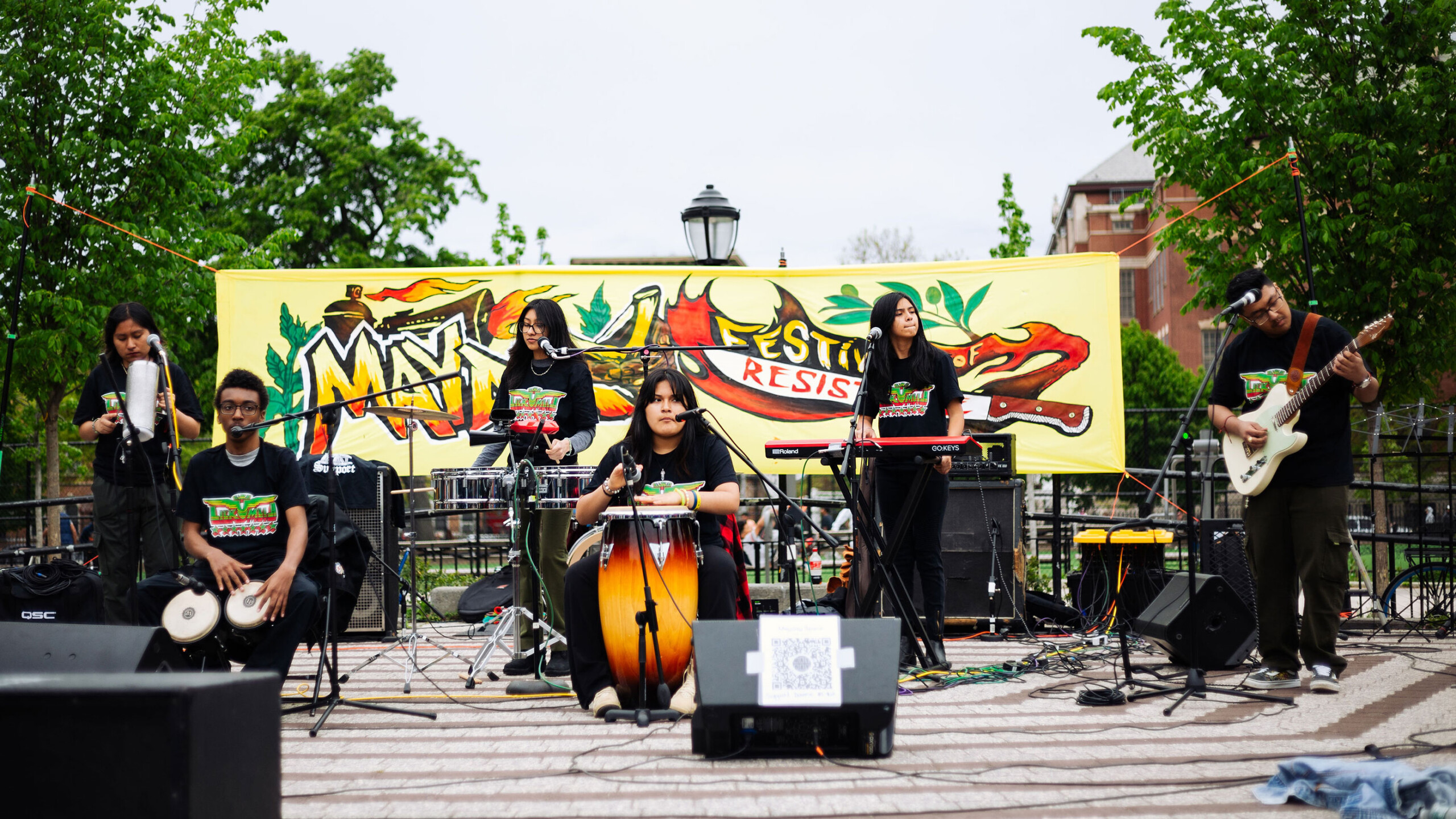 Band performing outdoors with colorful May Day Festival of Resistance banner in the background.