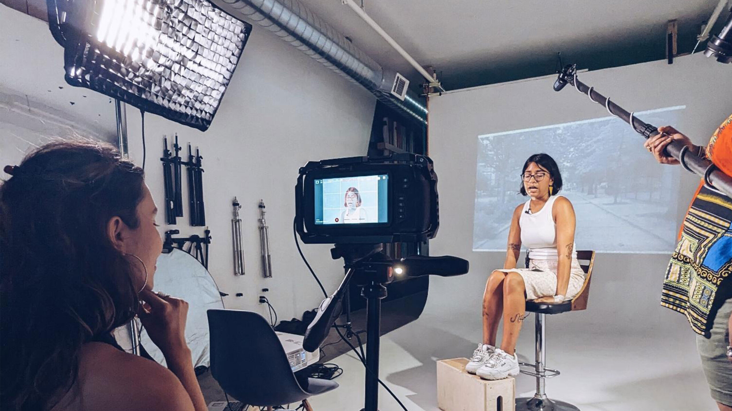A person is seated on a stool facing a camera during a filming session. The individual, wearing glasses and dressed in a light-colored sleeveless outfit with athletic...