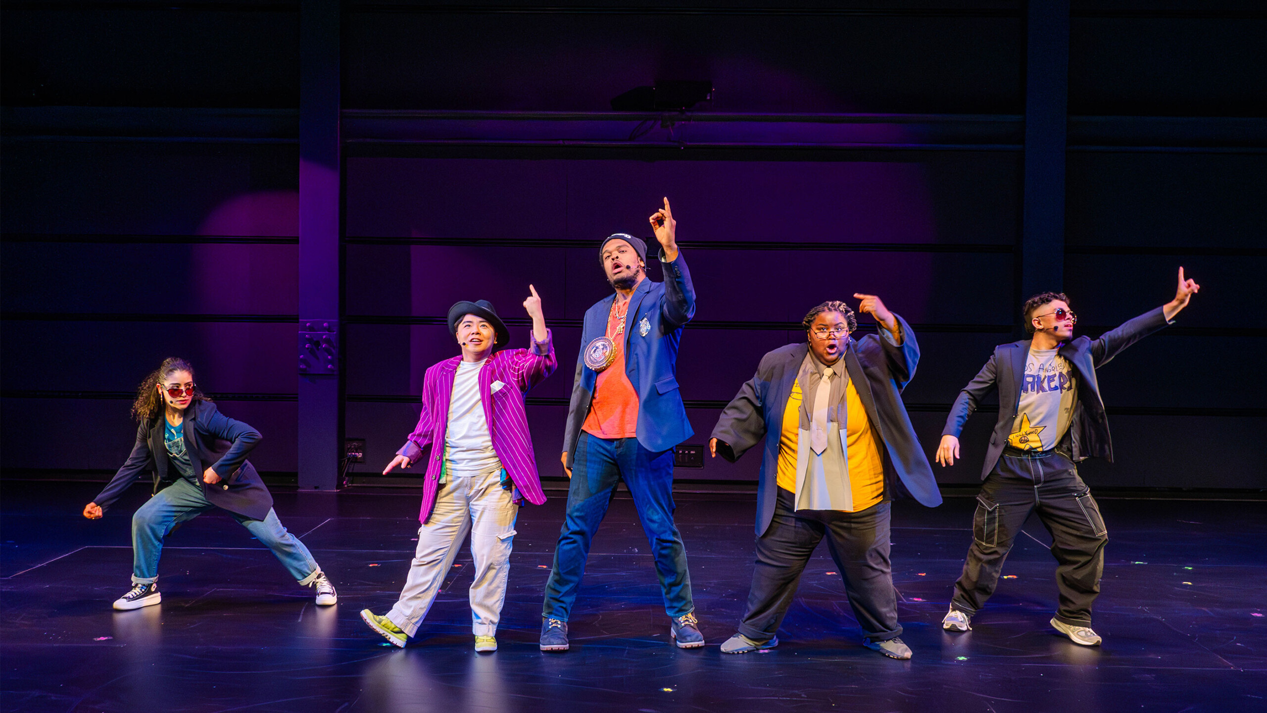 Five performers are dynamically posed on stage, each in a distinct and expressive stance. They all wear vibrant and eclectic outfits featuring blazers, sunglasses,...