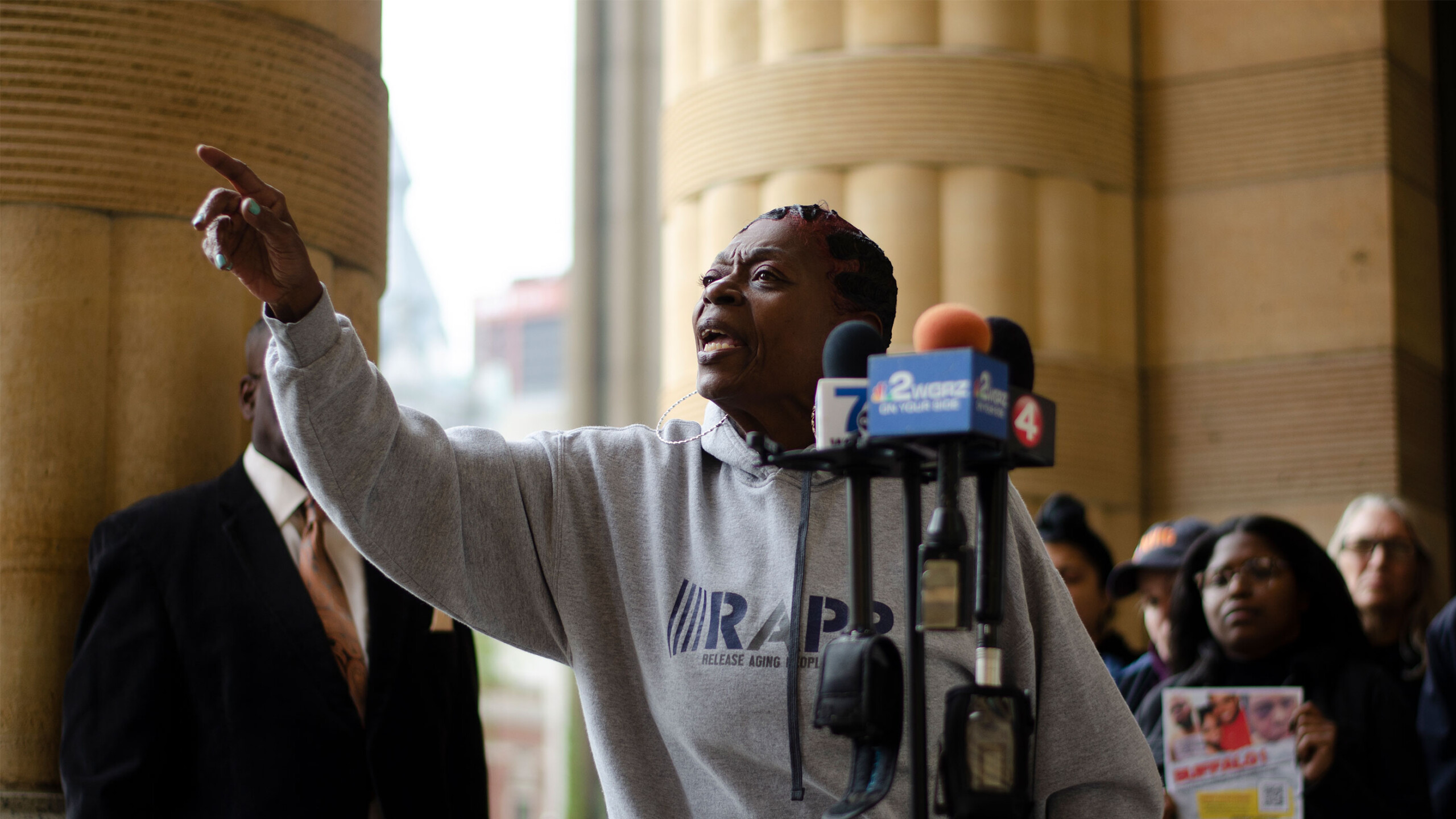 A person passionately addresses a crowd while standing in front of a row of microphones placed on a stand. Wearing a gray hoodie with a design or text on it, the...