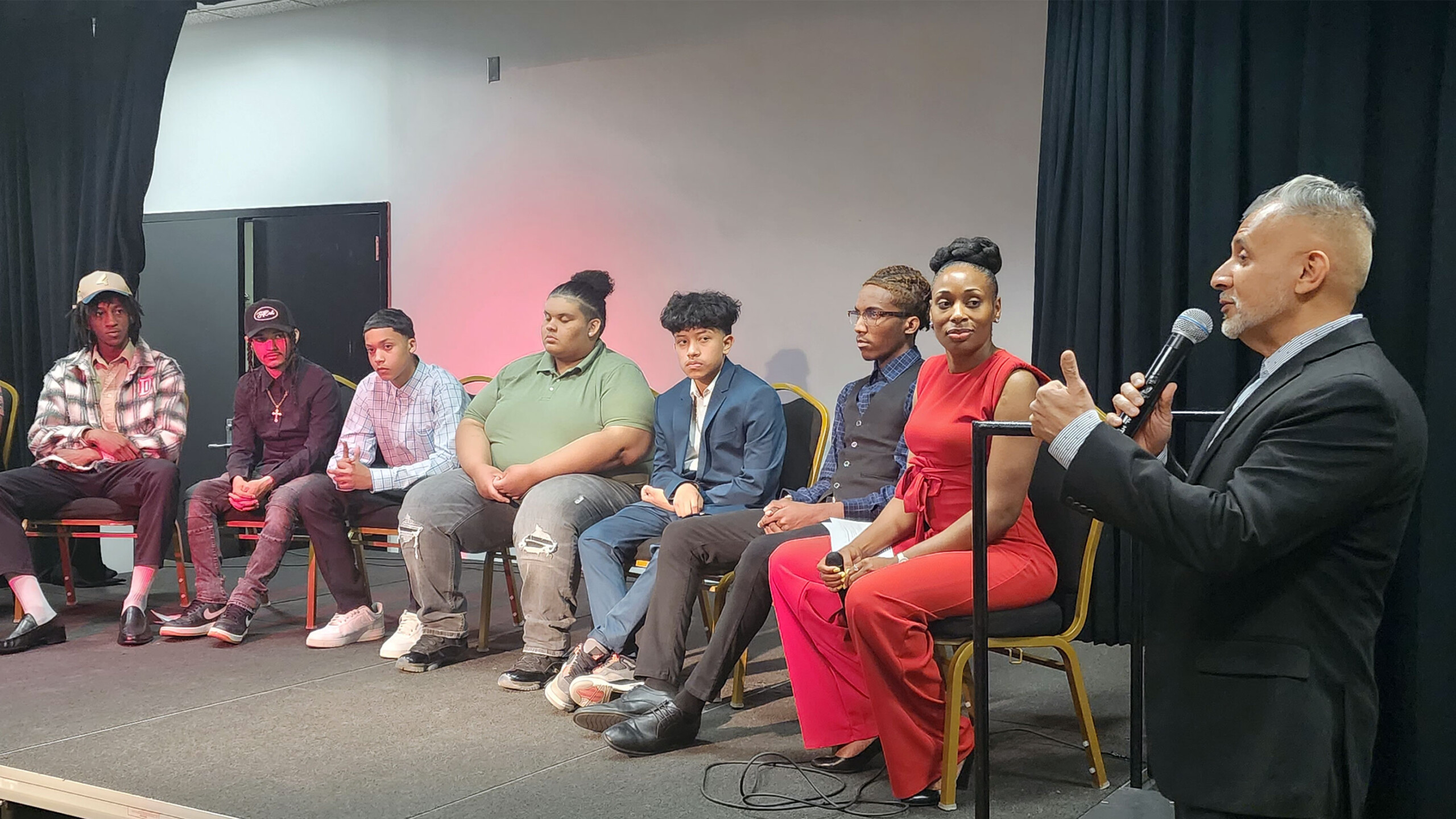 A group of seven individuals seated in a row on a stage, with one person standing to the right holding a microphone, suggesting they may be moderating or speaking...