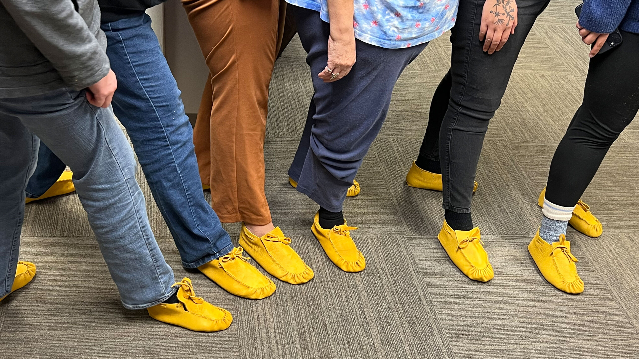 A group of people standing in a row, each wearing brightly colored yellow moccasins. The individuals are dressed in various casual outfits, including blue jeans,...