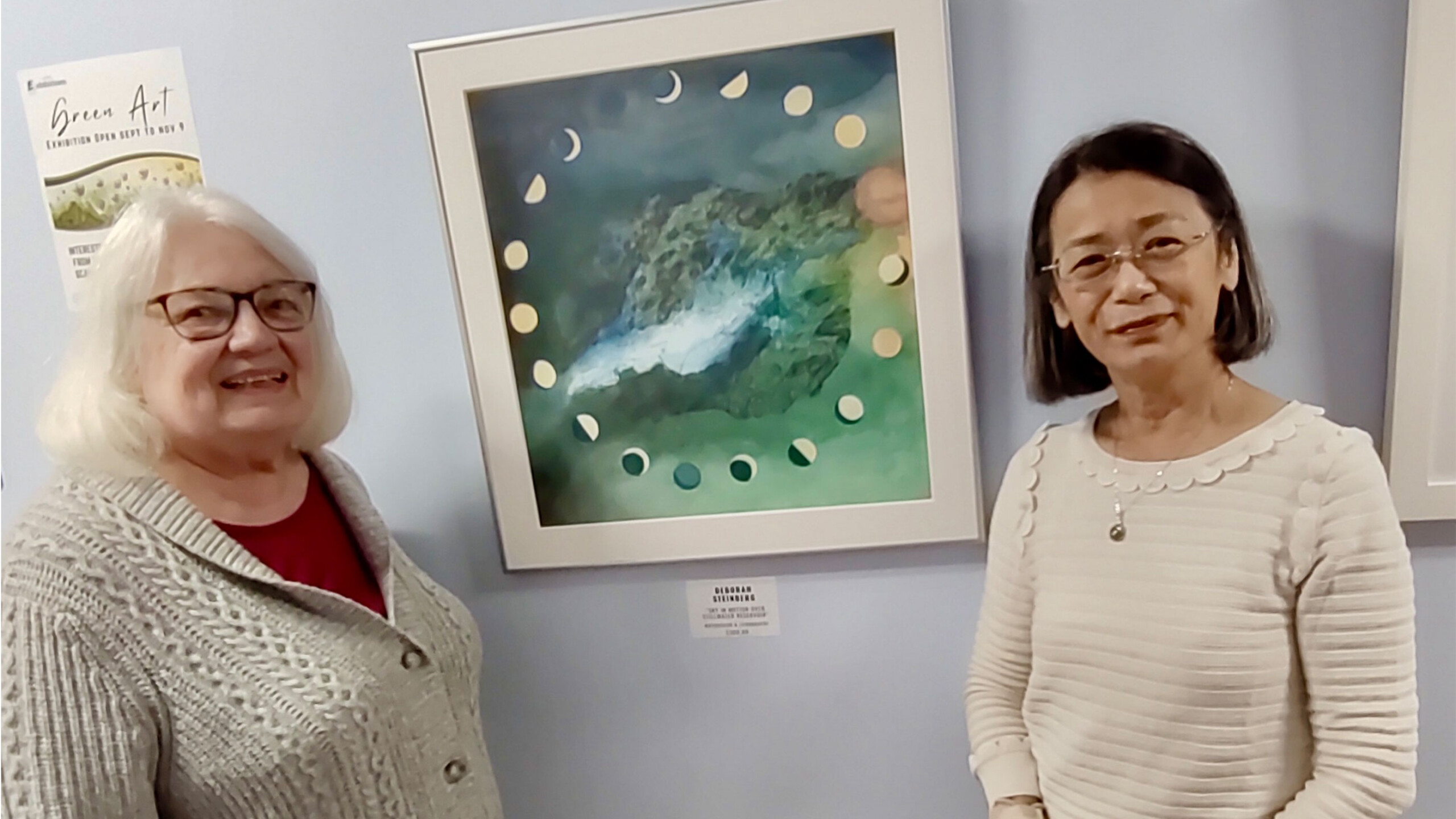 Two women are standing beside a piece of framed artwork displayed on a light blue wall. The woman on the left has gray hair and is wearing a light gray cardigan...