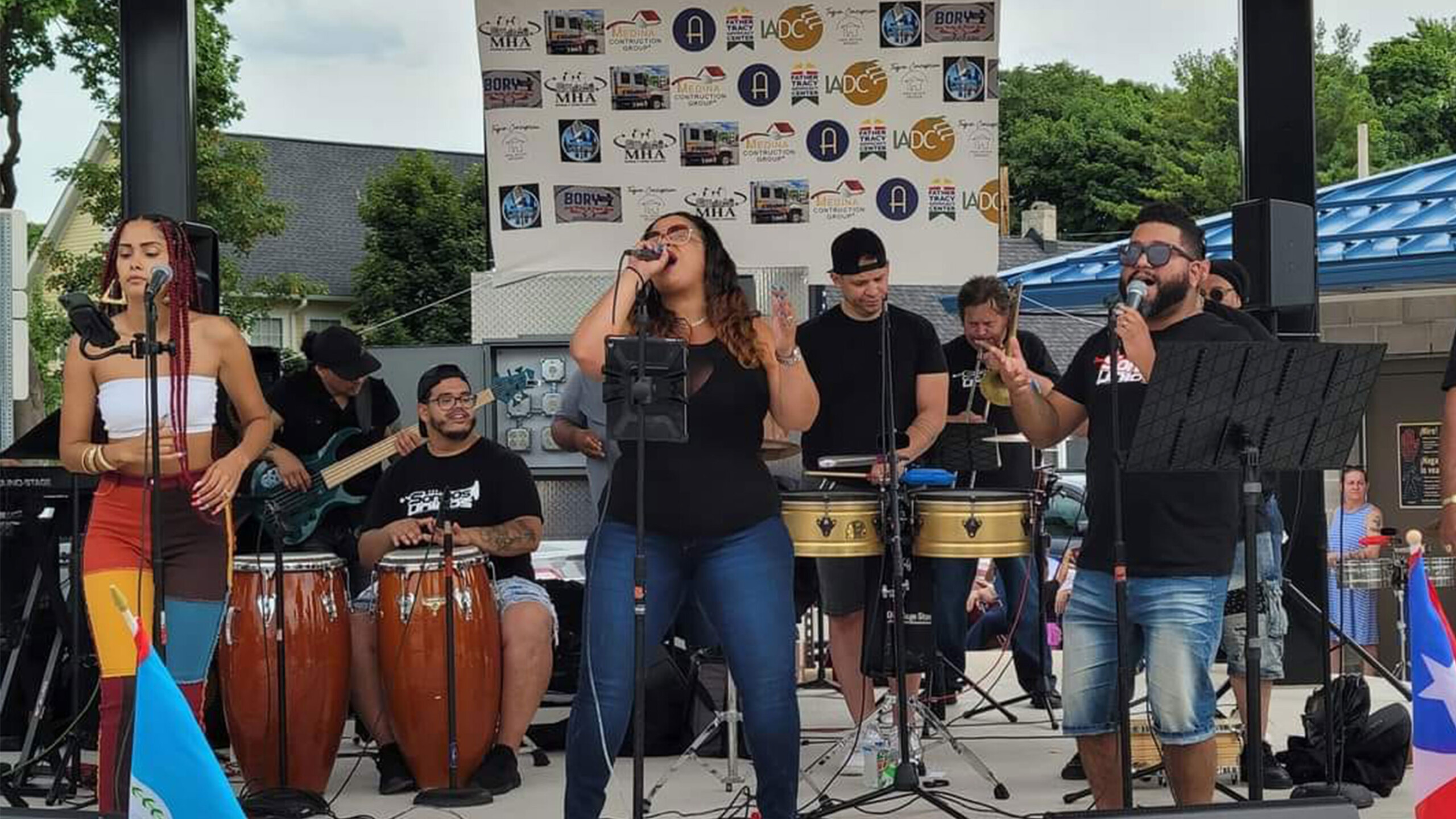 A lively band is performing on an outdoor stage, featuring a group of musicians passionately engaged in a musical performance. Two vocalists stand at the front,...