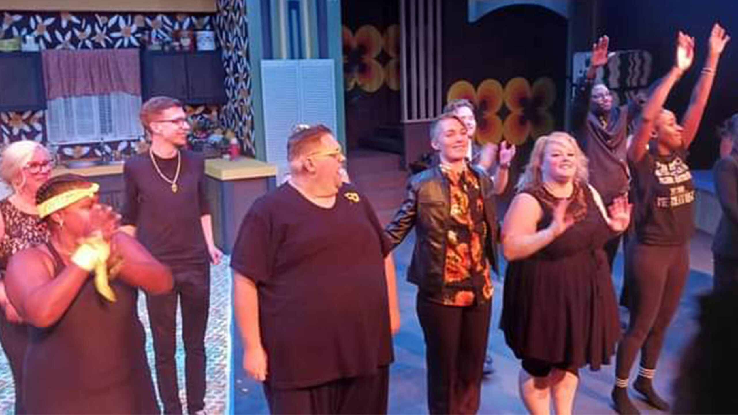 A group of performers stands on stage in a casual setting, likely having just finished a performance. The stage backdrop features retro-style decor with floral...