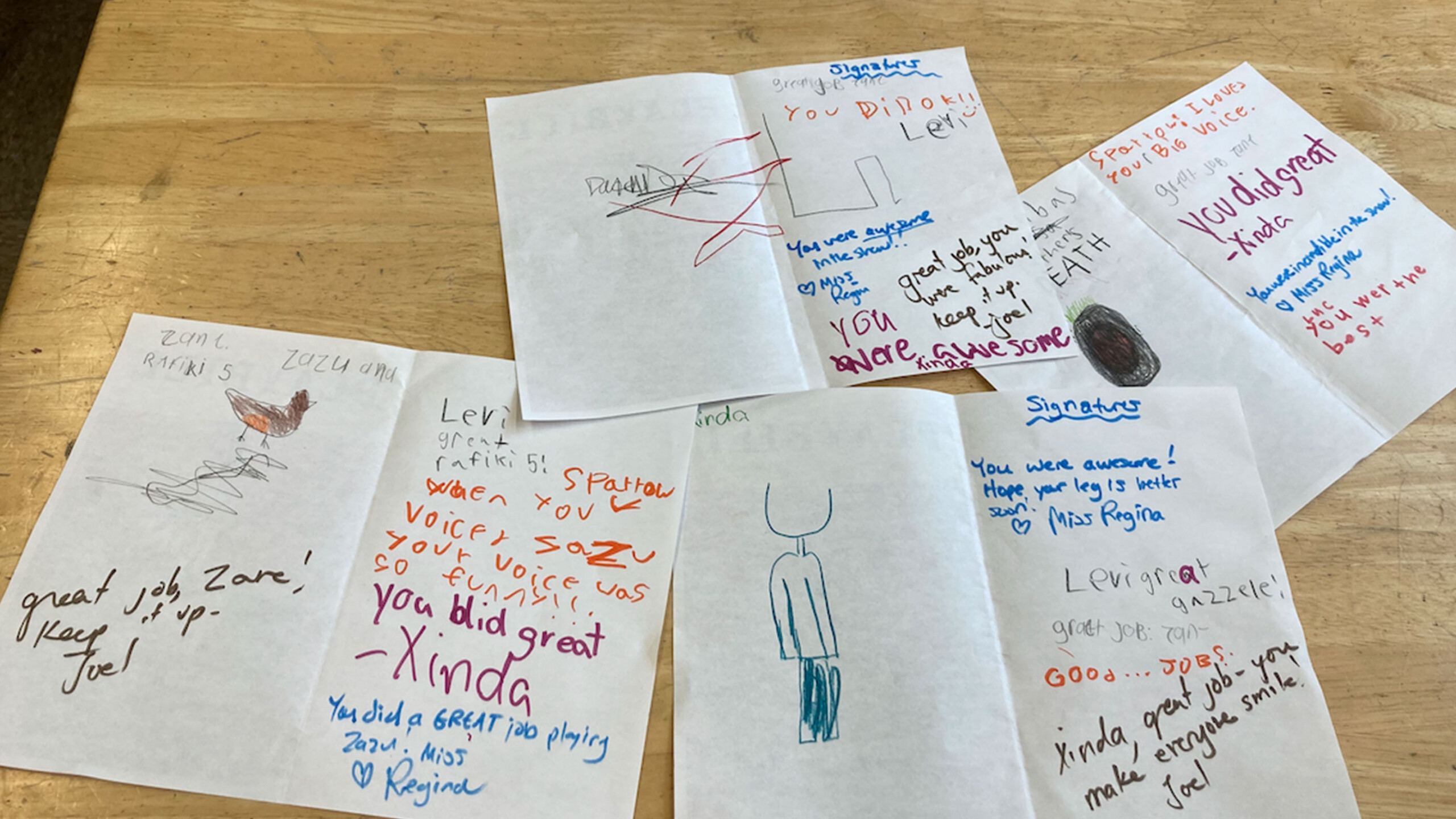 Several sheets of paper are laid out on a wooden table, each containing colorful drawings and handwritten messages. The papers include various artistic expressions...