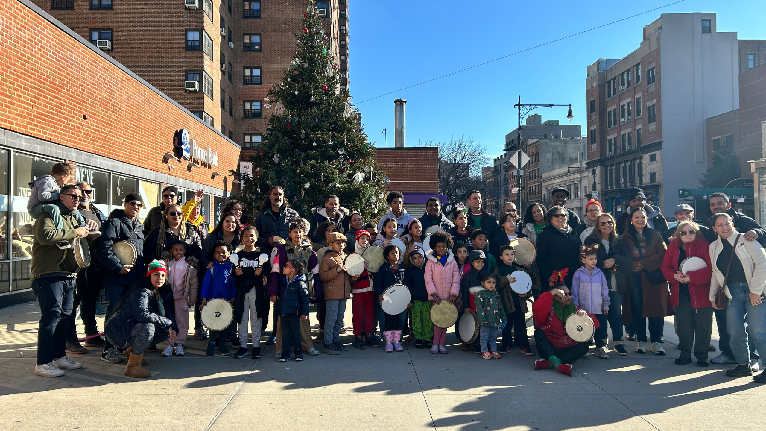 A large group of people, consisting of both adults and children, gathered in an urban setting for a festive occasion. In the background, a decorated Christmas tree...