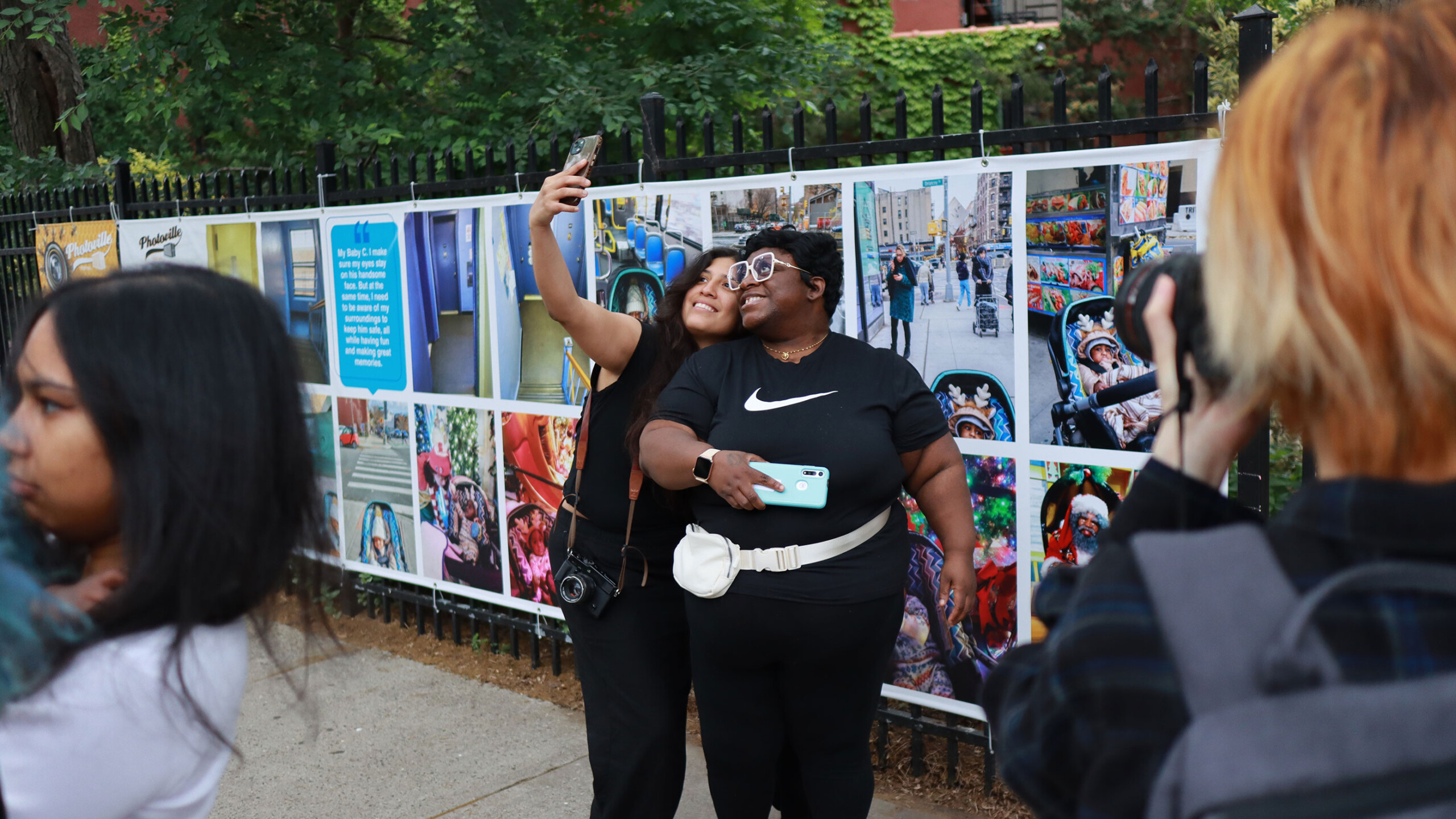 Two people pose happily for a selfie in front of a wall displaying a series of colorful and vibrant photographs. They stand close together, smiling broadly, with...