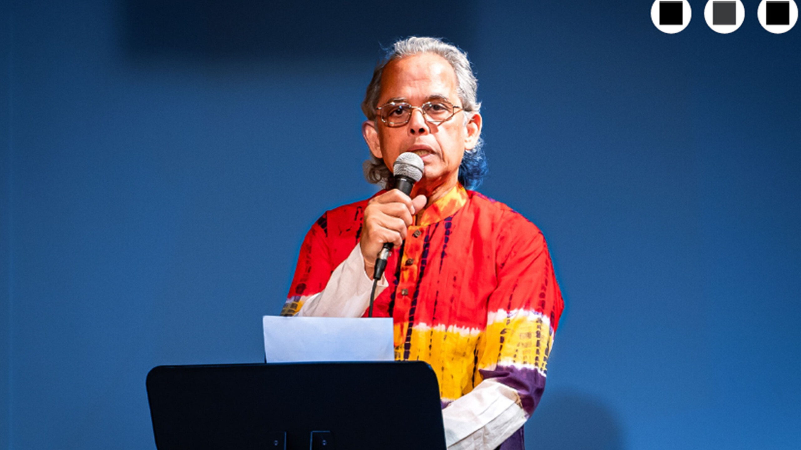Person in colorful shirt speaking into microphone with laptop on podium.