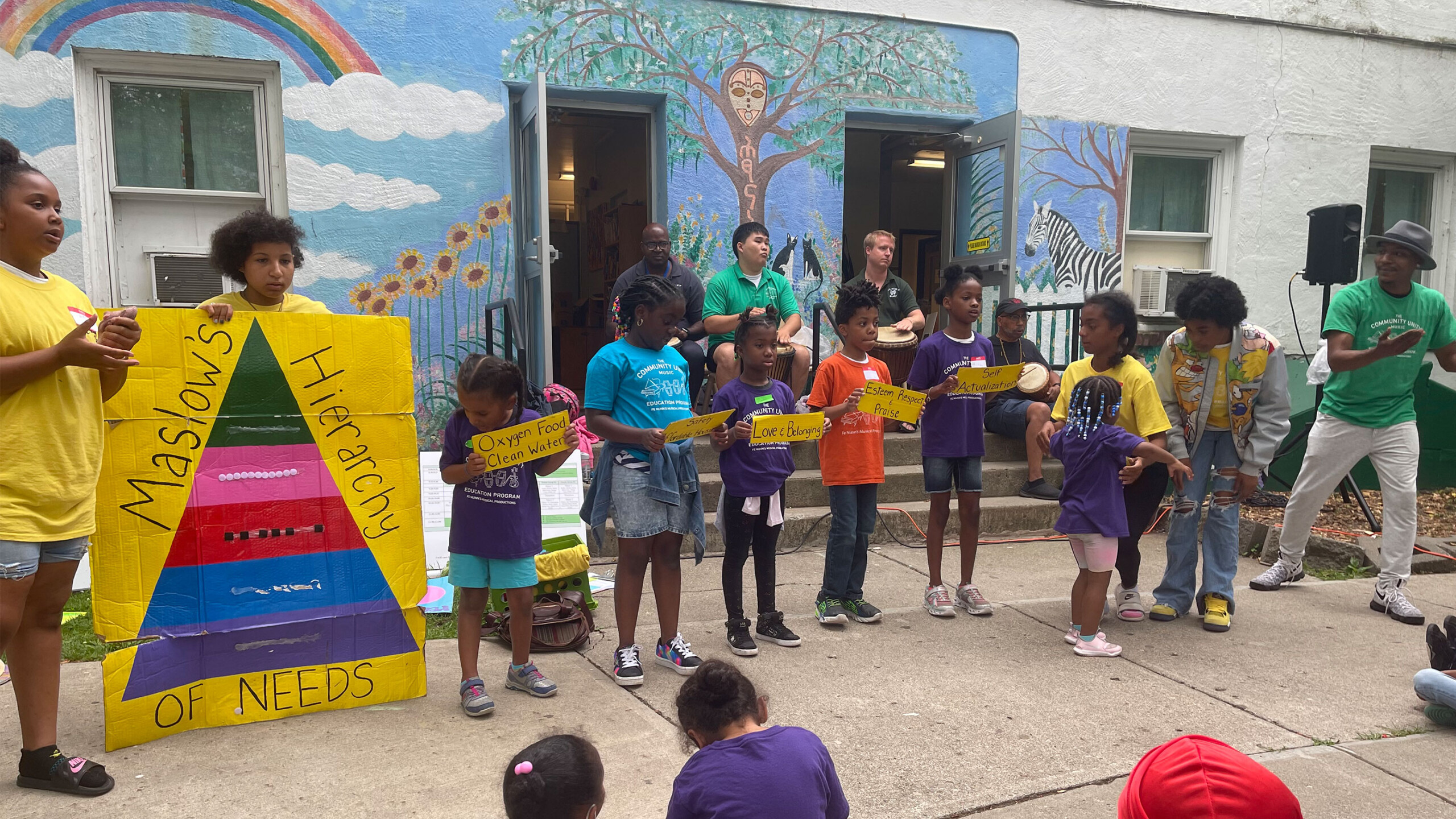 A group of children are participating in an educational presentation outdoors. They stand in a row holding signs that collectively form a message. Two of the children...