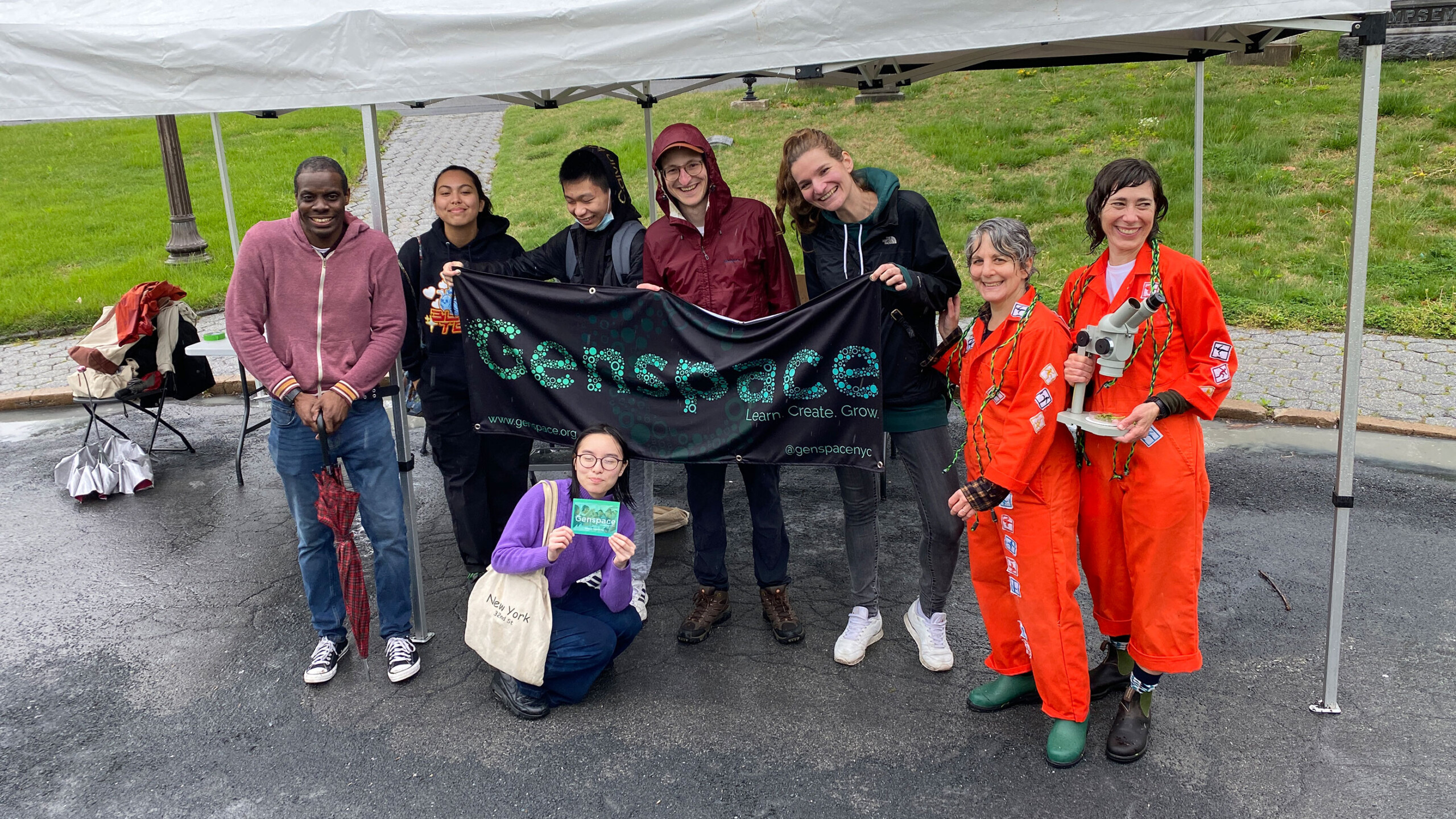 Group holding a Genspace banner at an outdoor event under a tent.