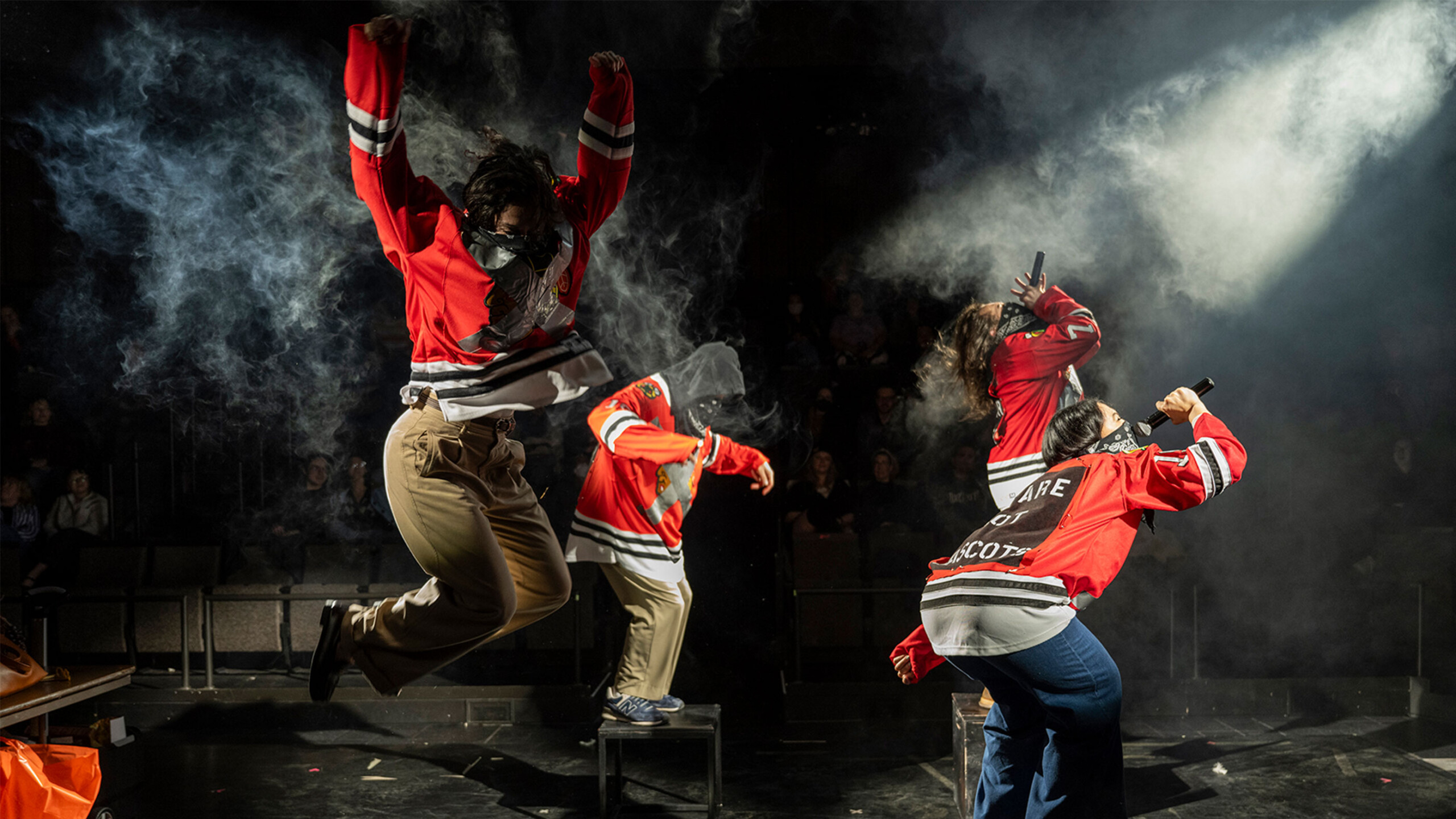Three performers on stage wear red sports jerseys, dynamically engaged in action. One performer is mid-air in a dramatic leap with arms raised, embodying energy...