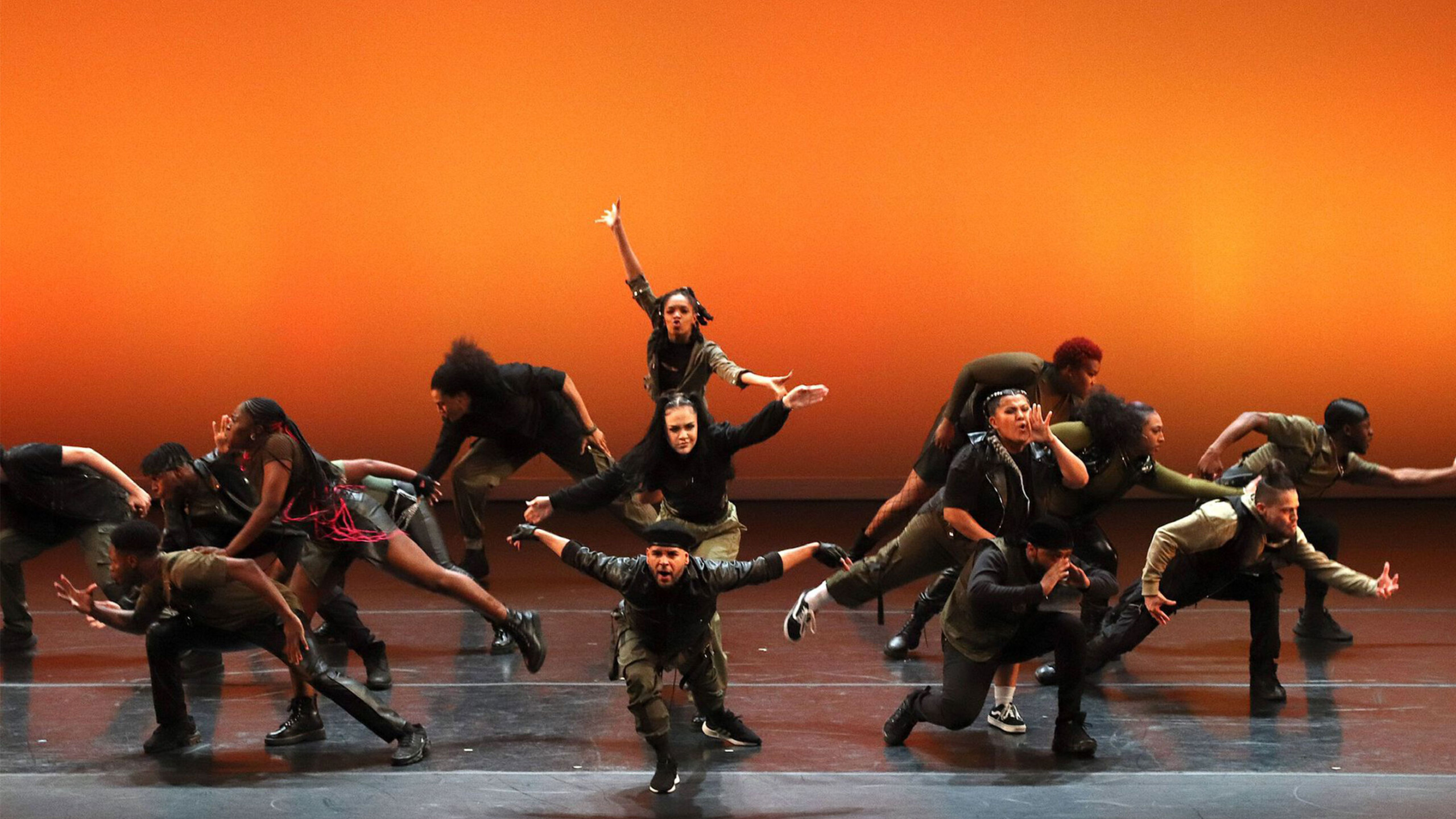 A dynamic group of dancers performing on stage against a warm, orange background. Various performers are captured in mid-motion, showcasing high energy and synchronized...