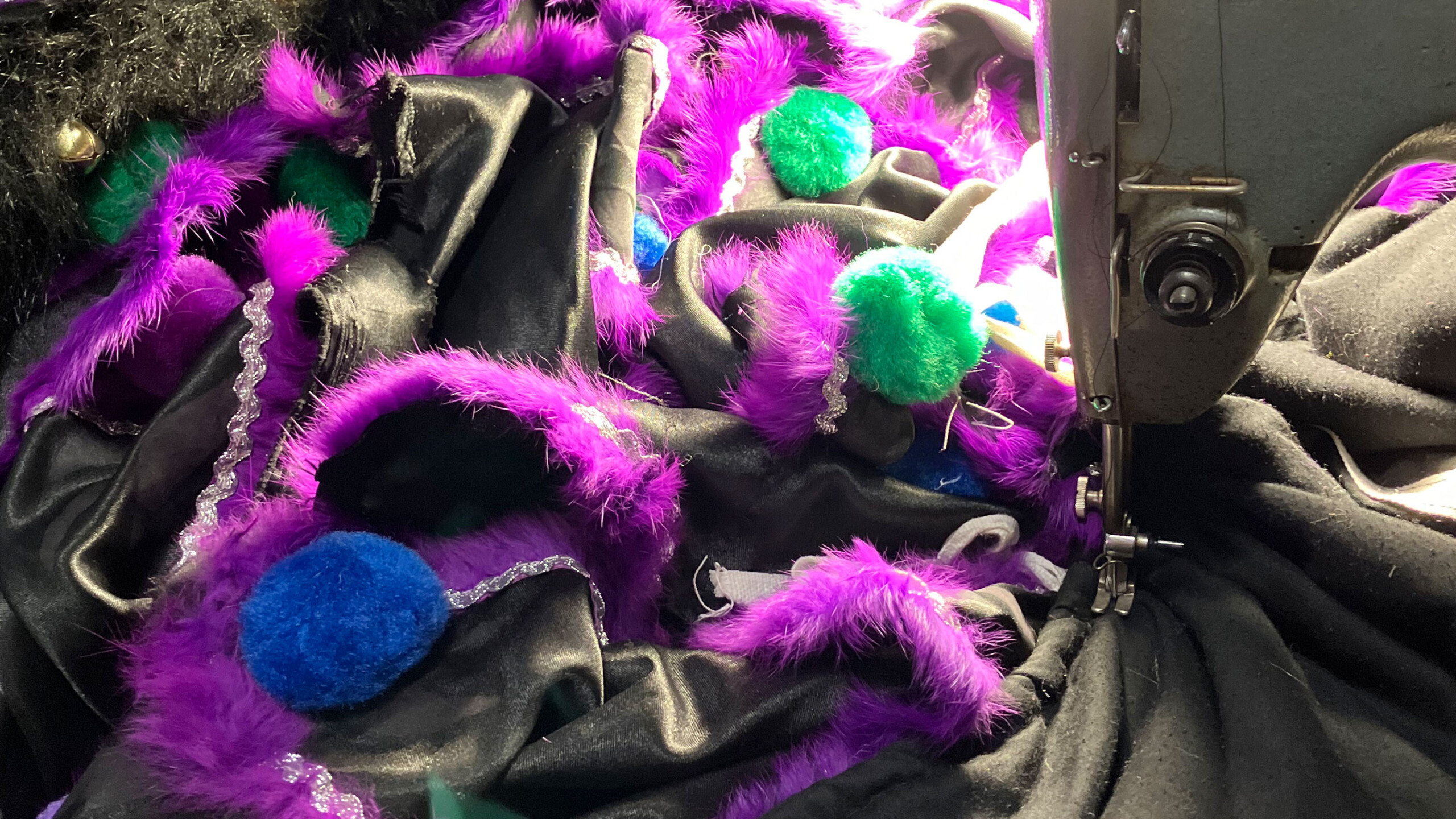 A colorful array of black and purple fuzzy fabric is draped around a sewing machine. Bright purple feathers and blue and green pom-poms embellish the fabric, alongside...