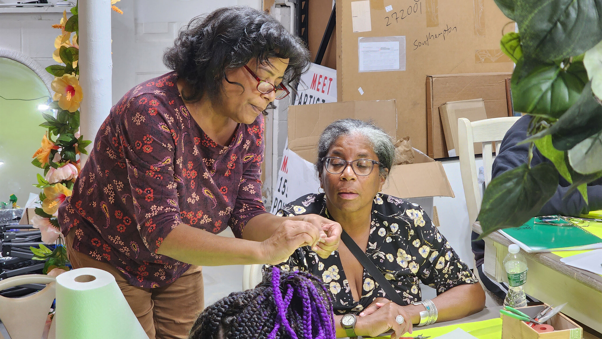 Two individuals in a cluttered office space with cardboard boxes and artificial flowers.