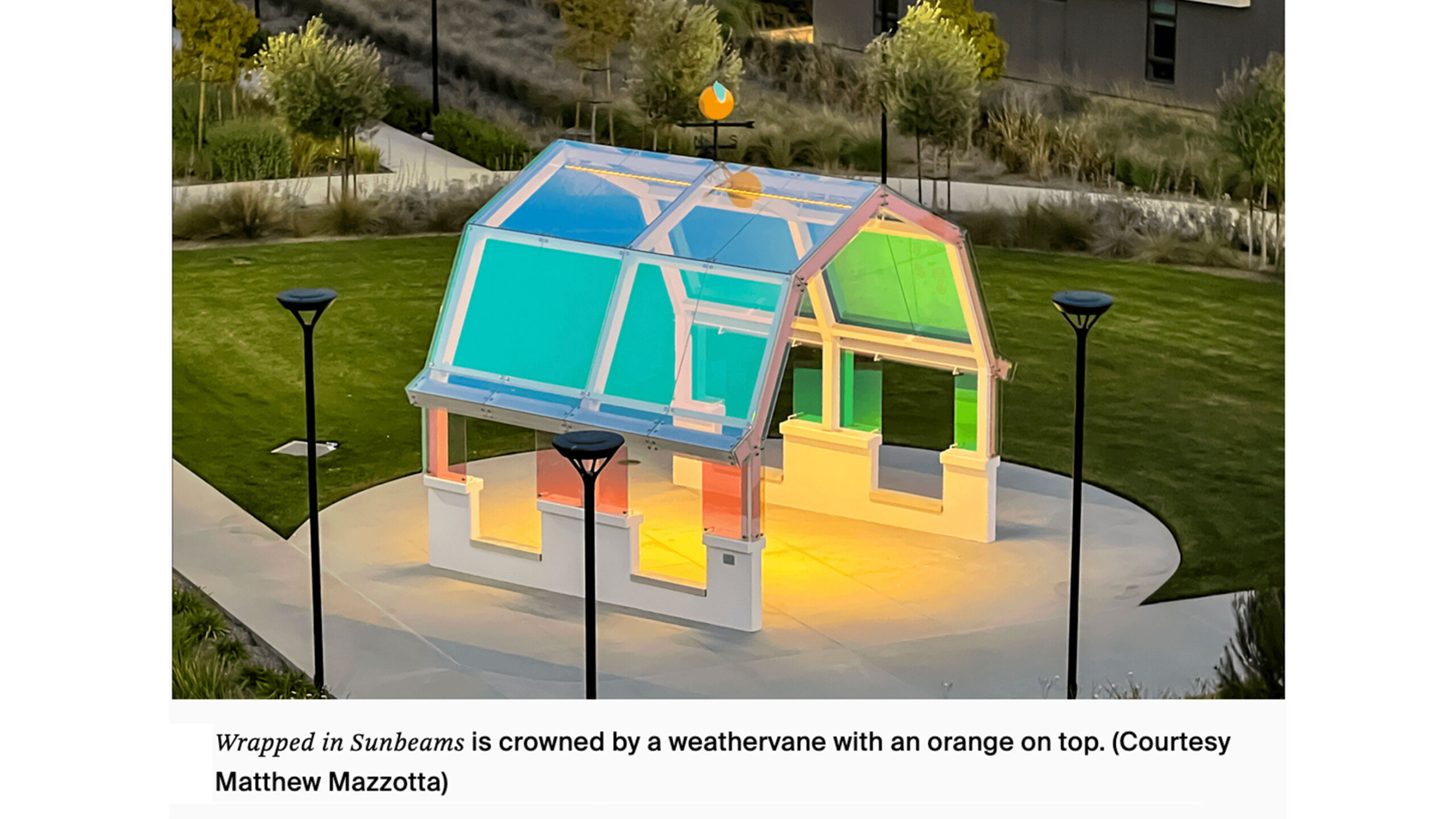 A small, transparent structure with a gabled roof stands prominently in an open landscaped area. The pavilion is designed with colorful panels, showcasing hues...
