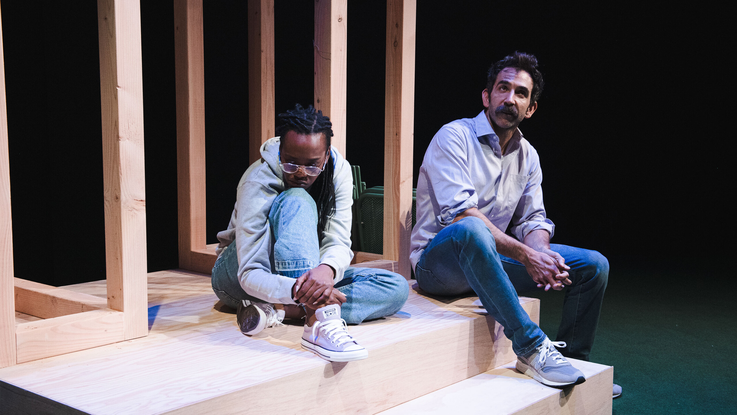 Two individuals sit on a wooden stage, framed by vertical wooden beams. One person wears a light-colored hoodie and jeans, with their arms wrapped around their...