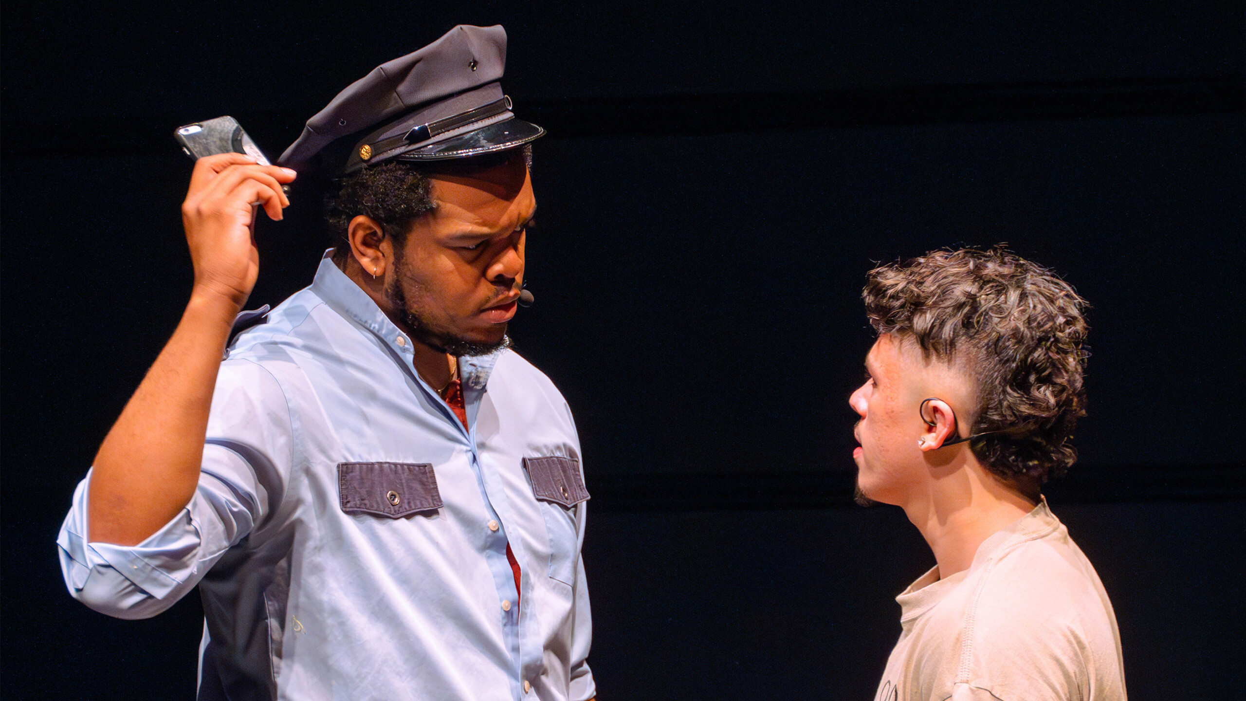 Two actors are in the middle of an intense scene on stage. The actor on the left wears a blue uniform shirt with dark patches on the chest and a matching cap, suggesting...