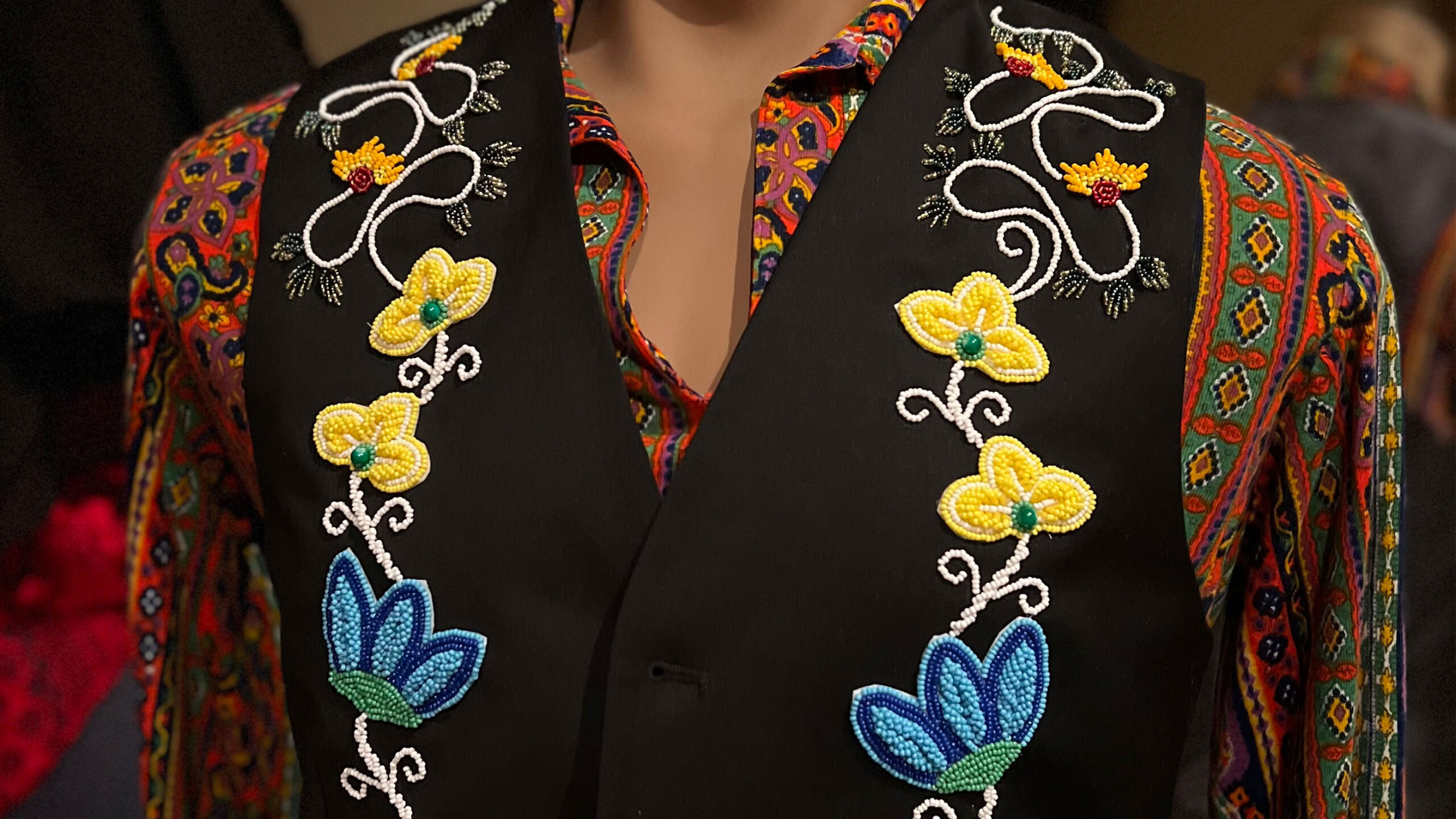 A vibrant embroidered vest worn over a colorful patterned shirt. The vest is black and adorned with intricate floral embroidery featuring yellow and blue flowers...