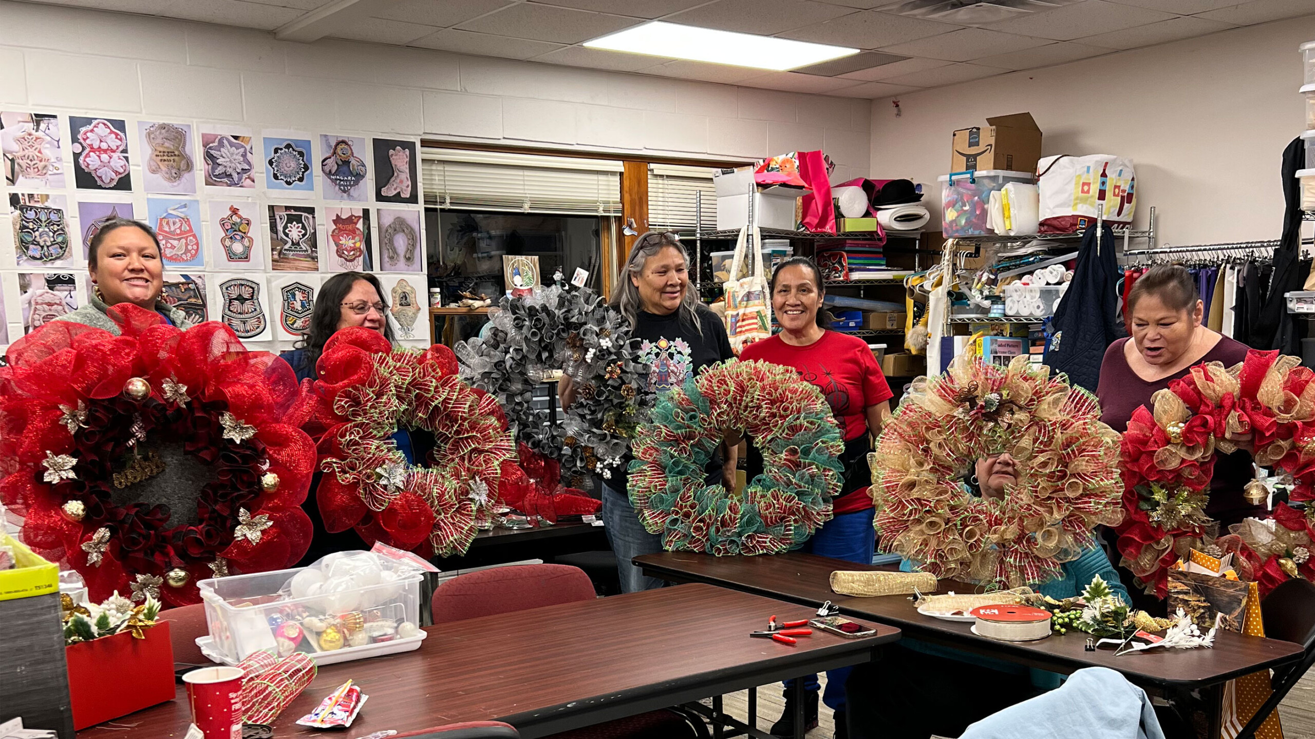 A group of five people standing in a workspace, proudly holding large, decorative holiday wreaths. The wreaths are diverse in color, featuring red, green, and gold...