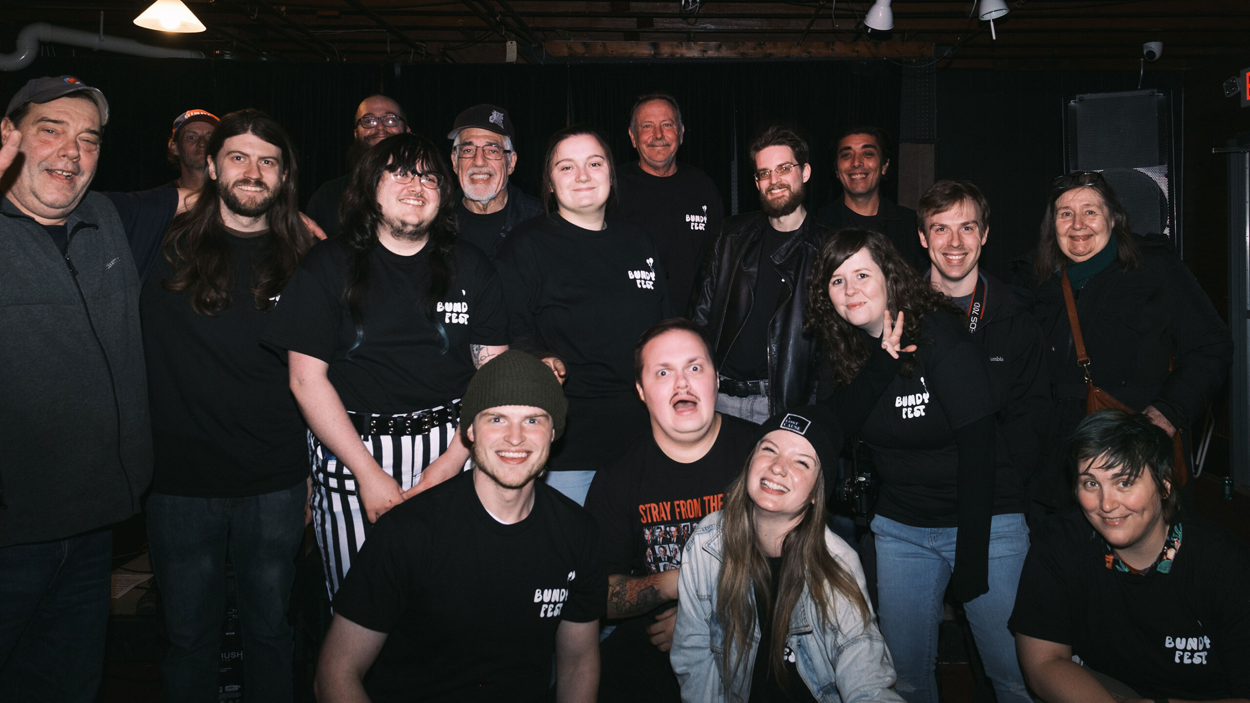 A diverse group of individuals poses happily together in a dimly lit indoor setting. There are around fifteen people, with some sitting in the front and others...