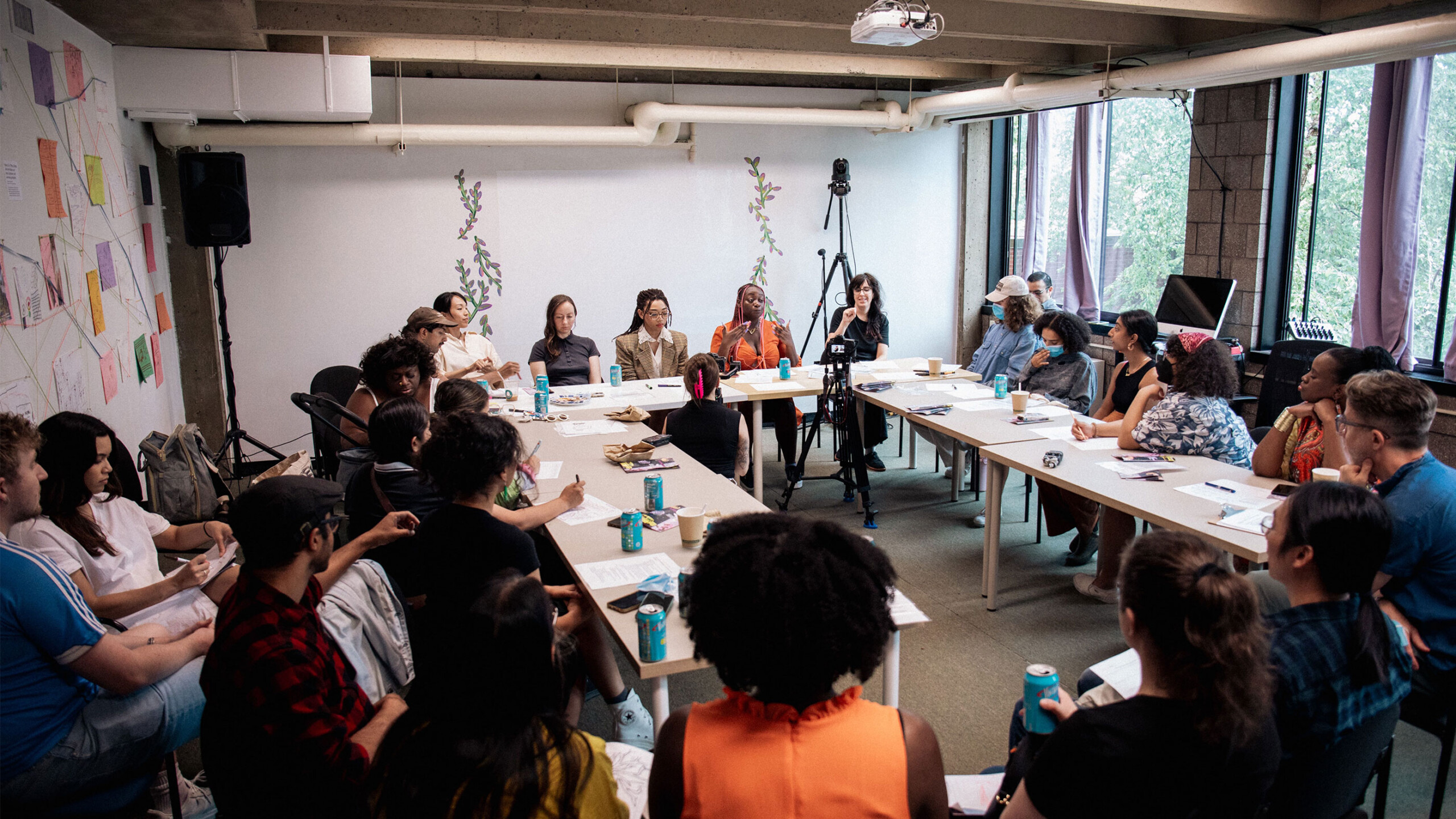 A diverse group of people sitting around two large connected tables in a bright, open room. Windows on the right wall let in natural light, providing a view of...