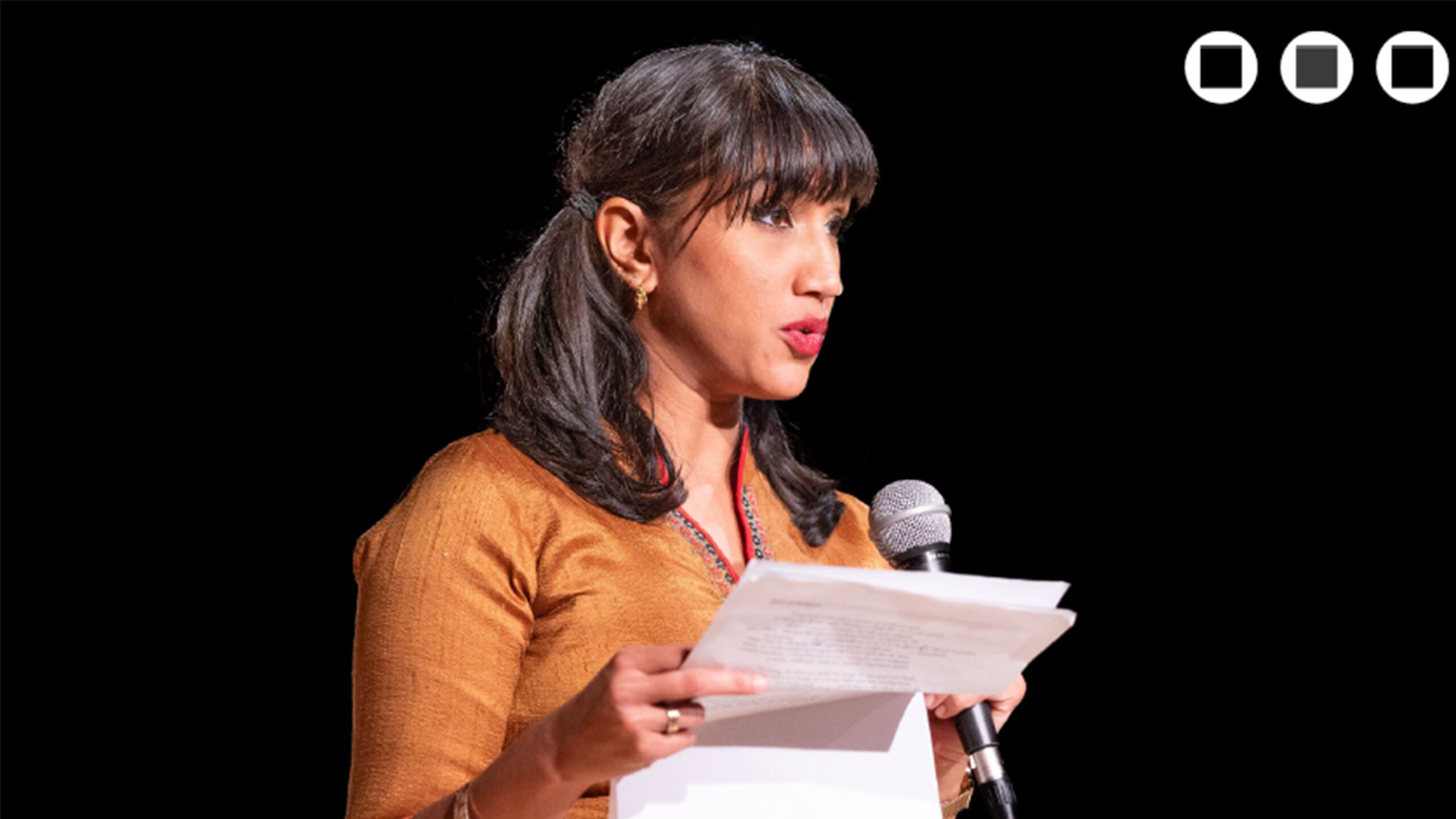 Person reading a paper at a microphone on a dark stage.