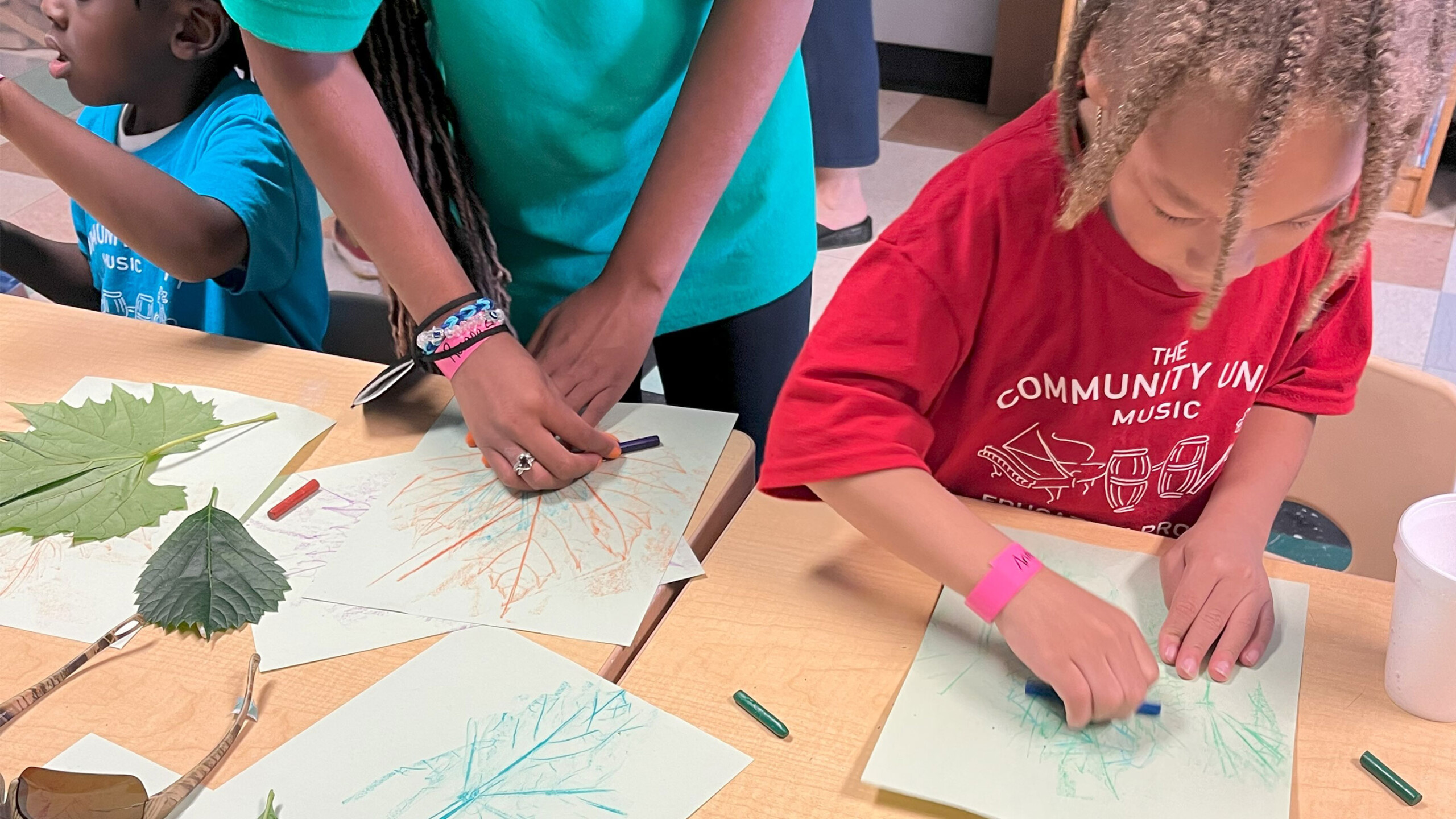 Children and an adult are engaged in a leaf rubbing activity at a classroom table. Each child has a sheet of paper placed over large, textured leaves and is using...