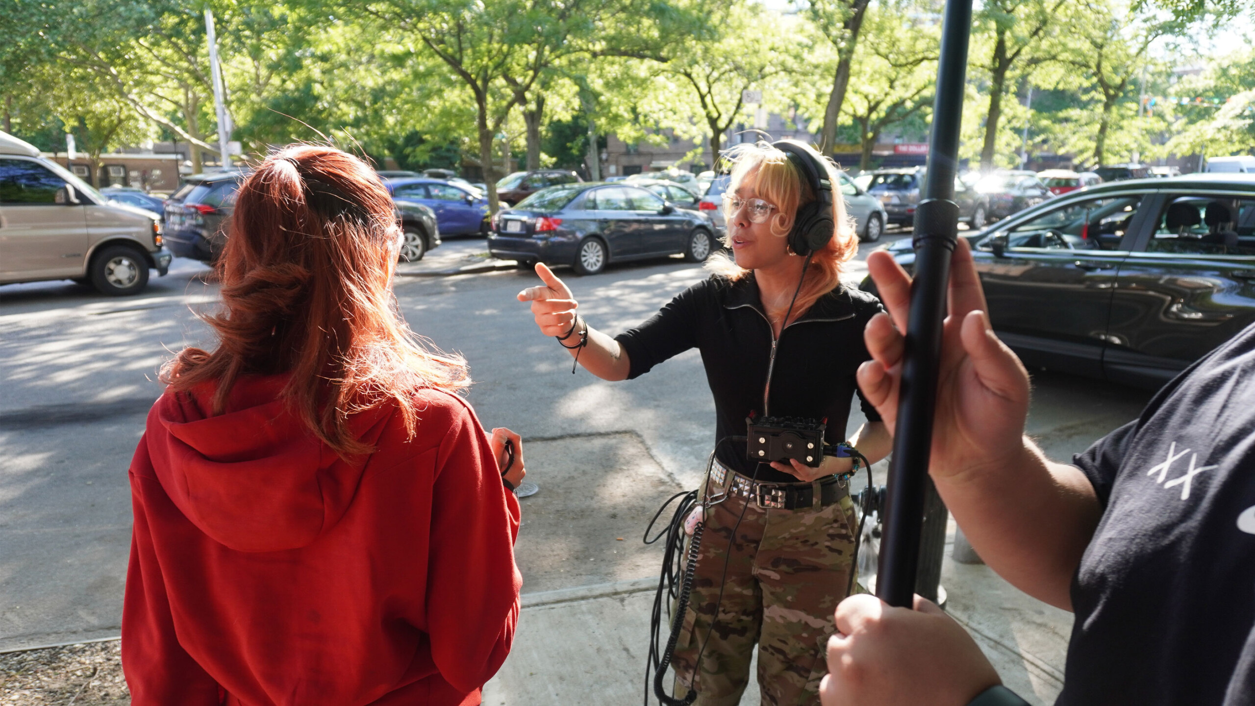 A woman wearing camouflage pants and a black top, equipped with headphones and holding a small audio mixer, is engaging in conversation with another person. This...