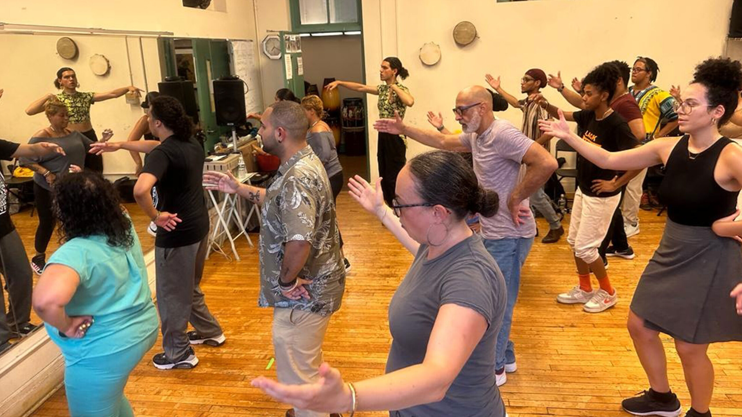 A diverse group of individuals is participating in a dance class held in a spacious room with wooden flooring. The participants, both men and women, are standing...