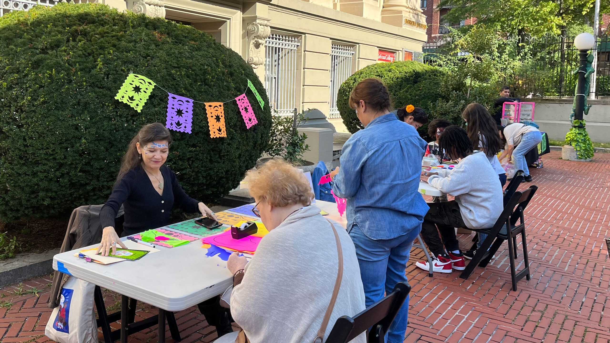 A group of people engage in a crafting activity at an outdoor event. They are seated at several tables arranged along a brick patio in front of a building. Colorful...