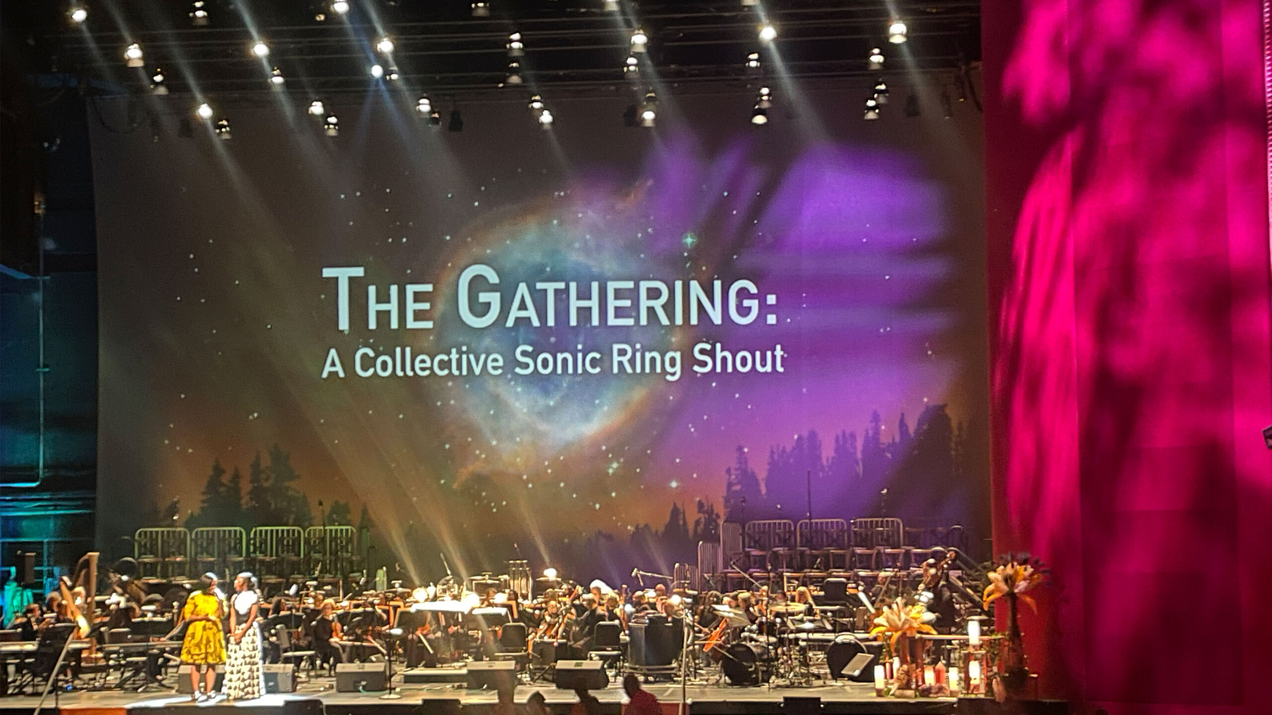 A grand stage with a prominent display of the event title 'The Gathering: A Collective Sonic Ring Shout' projected onto a backdrop featuring a cosmic and forest...