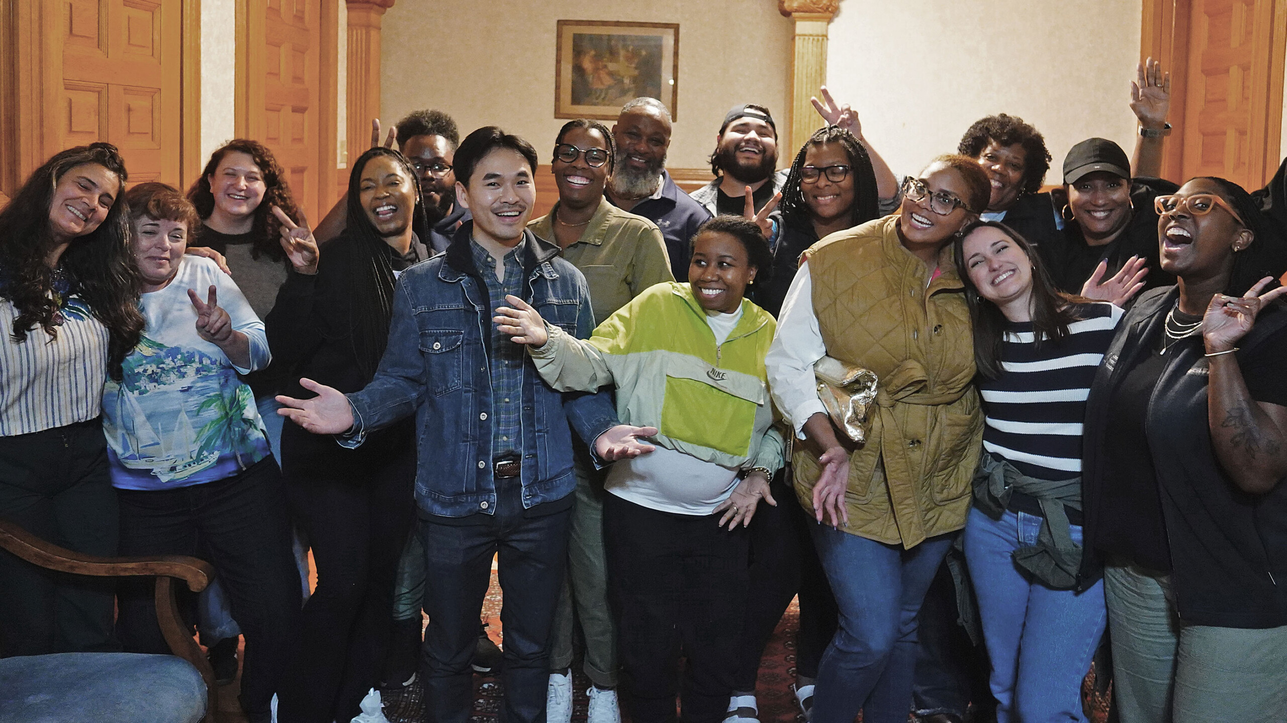 The image features a group of people posing together for a photo indoors. The participants are wearing casual clothing, including jeans and stylish footwear. They...