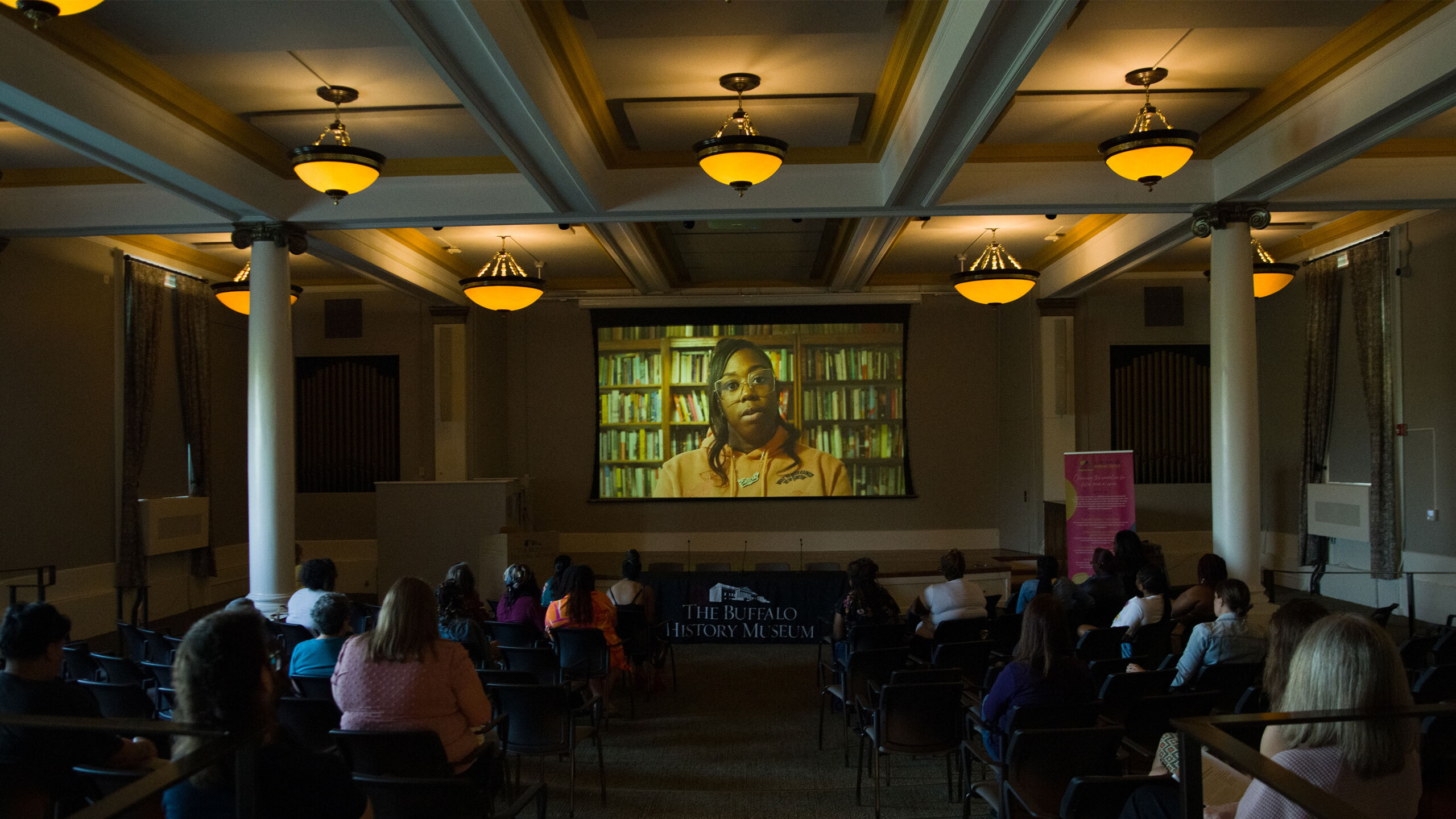 A group of people seated in a spacious room watching a film projected onto a large screen. The hall has an ornate ceiling with chandeliers providing ambient lighting....
