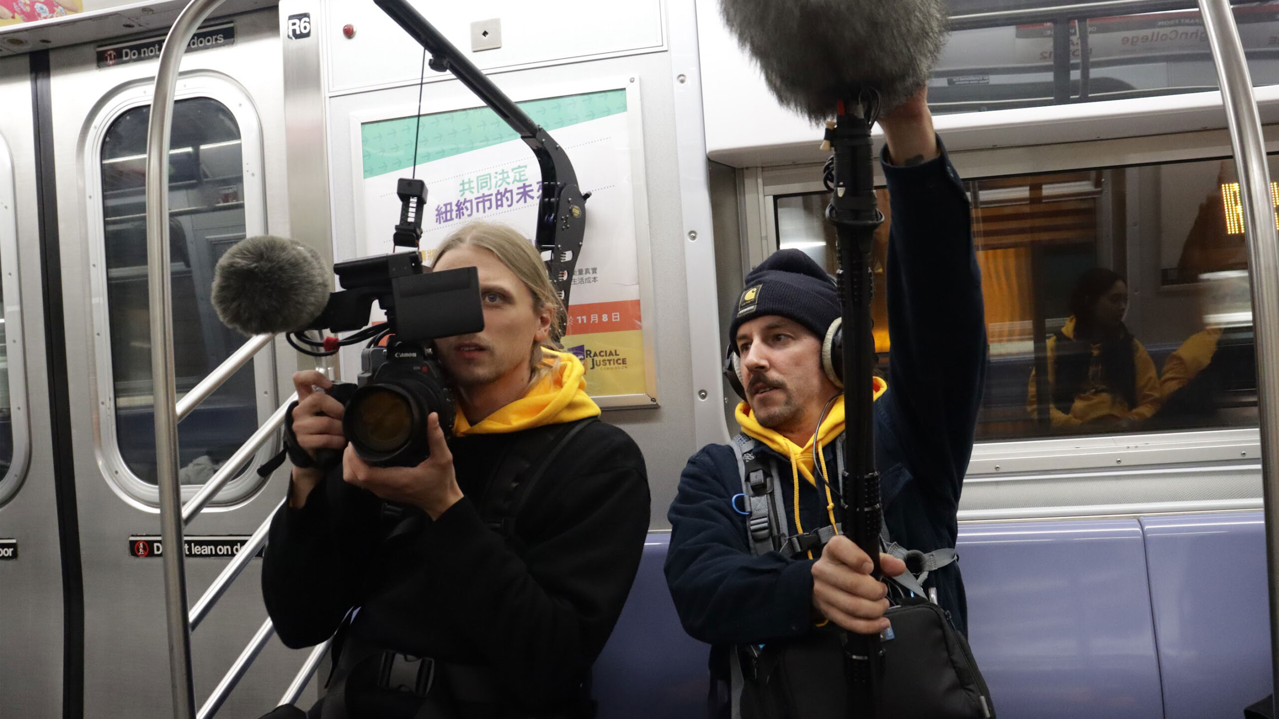 Two individuals are sitting inside a subway car engaged in filming. The person on the left is operating a professional video camera, equipped with a large microphone...