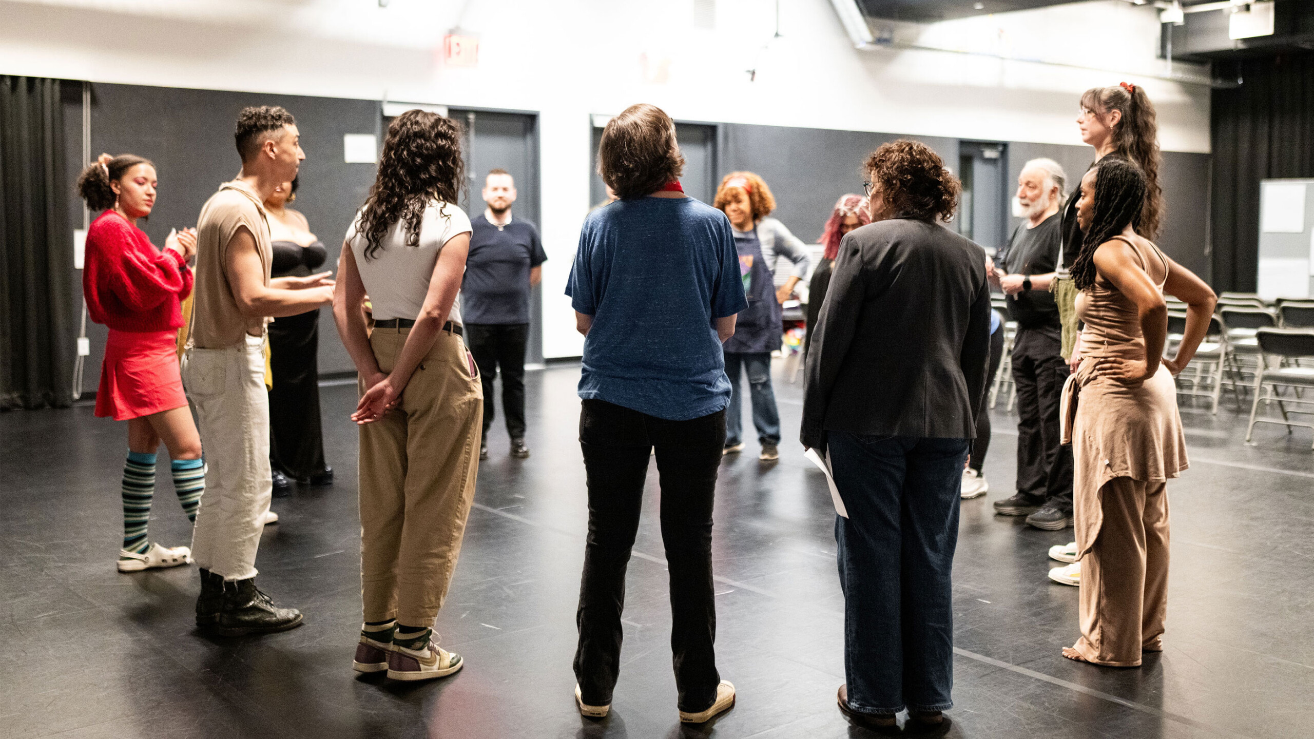 A diverse group of individuals stands in a circle inside a spacious dance studio. The participants are casually dressed, each facing inward towards the center of...