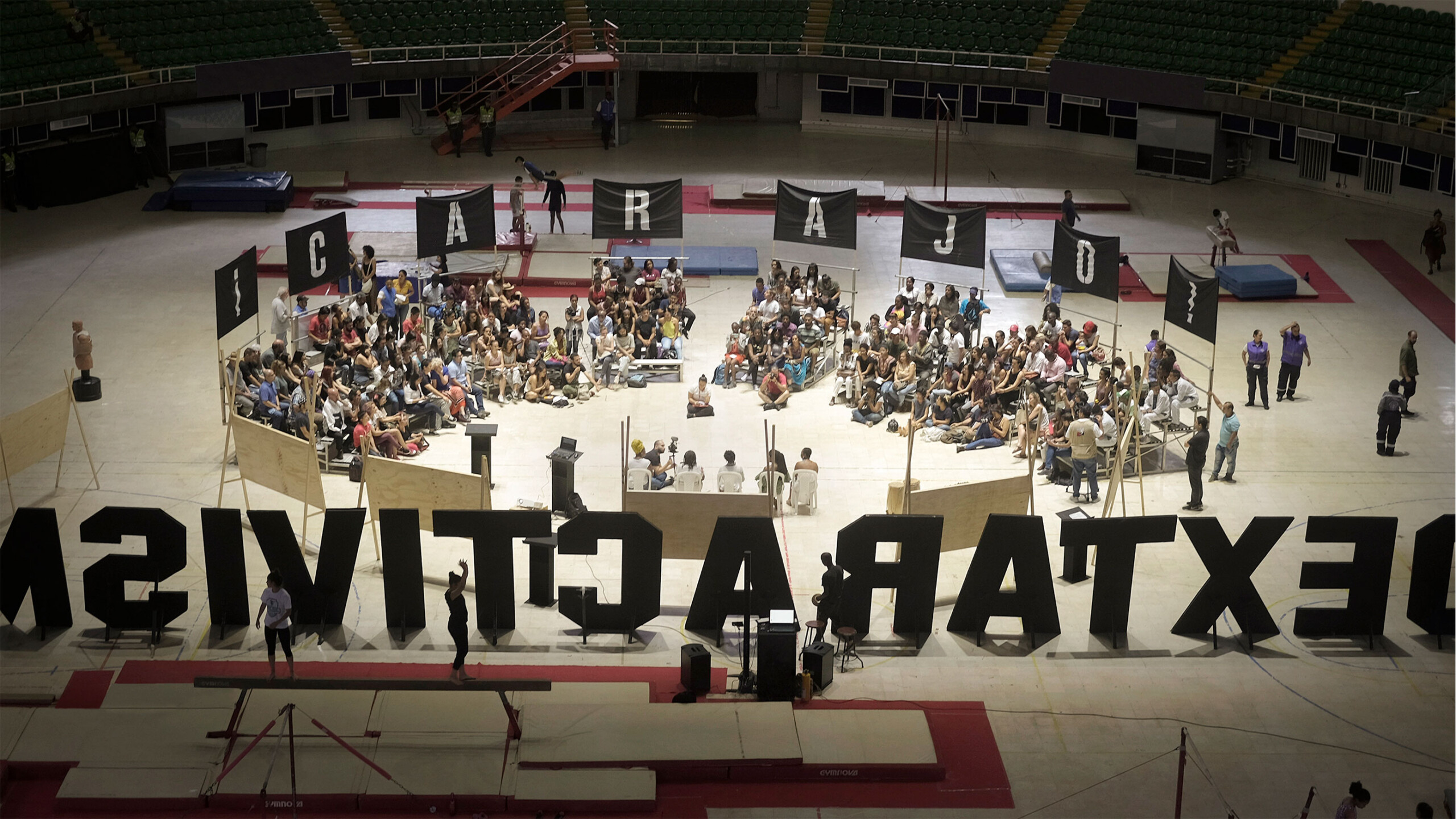 An indoor arena hosts a large community gathering, with a crowd seated on chairs arranged in a circular formation. Surrounding the circle on the floor are large...