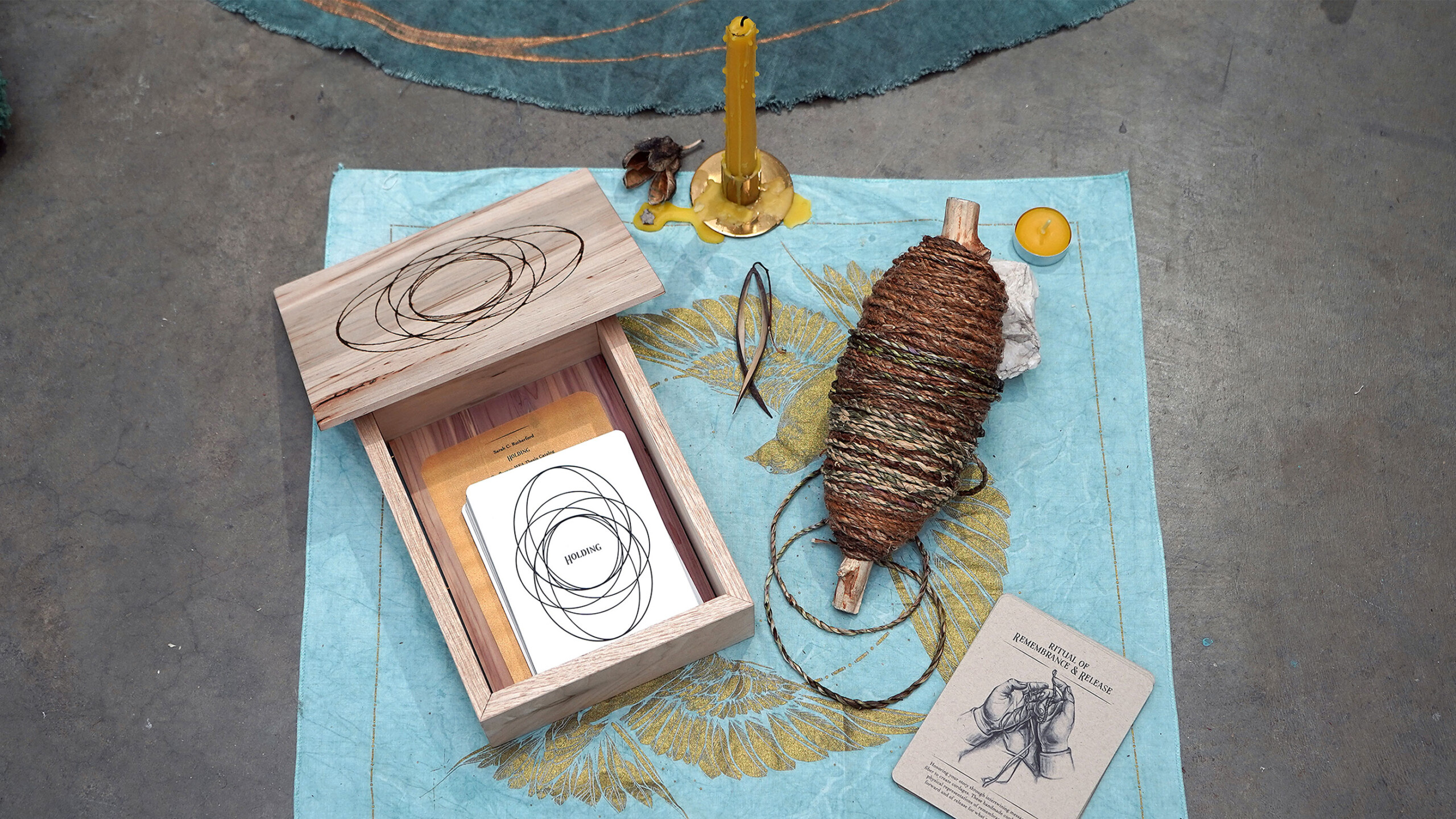 A collection of mystical objects arranged on a teal cloth, featuring a wooden box with spiral designs. Inside the box, a card with similar spiral patterns rests...