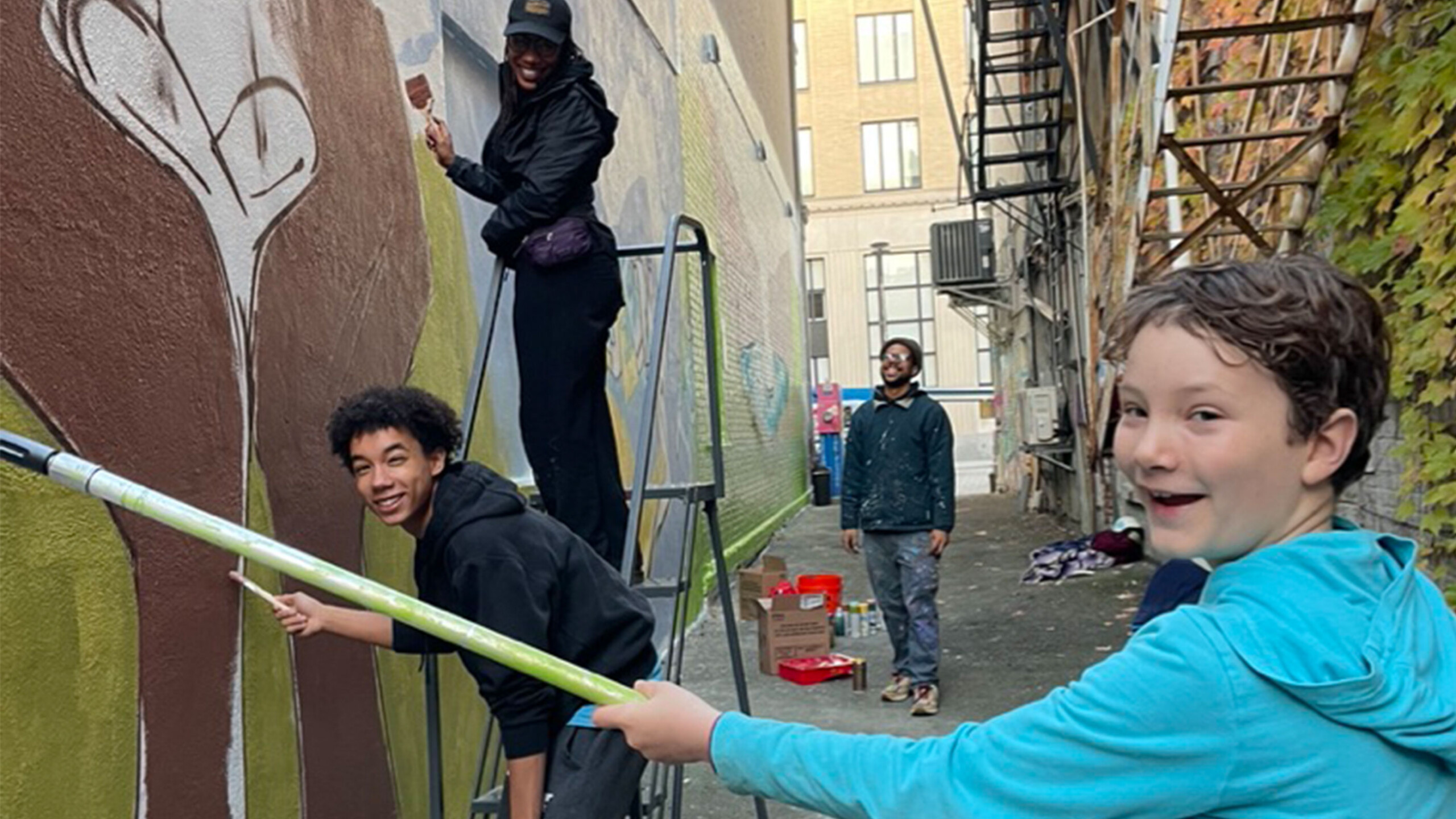 A group of people engaged in painting a mural on the wall of an alleyway. The alley walls are decorated with vibrant artwork, including nature and abstract designs....