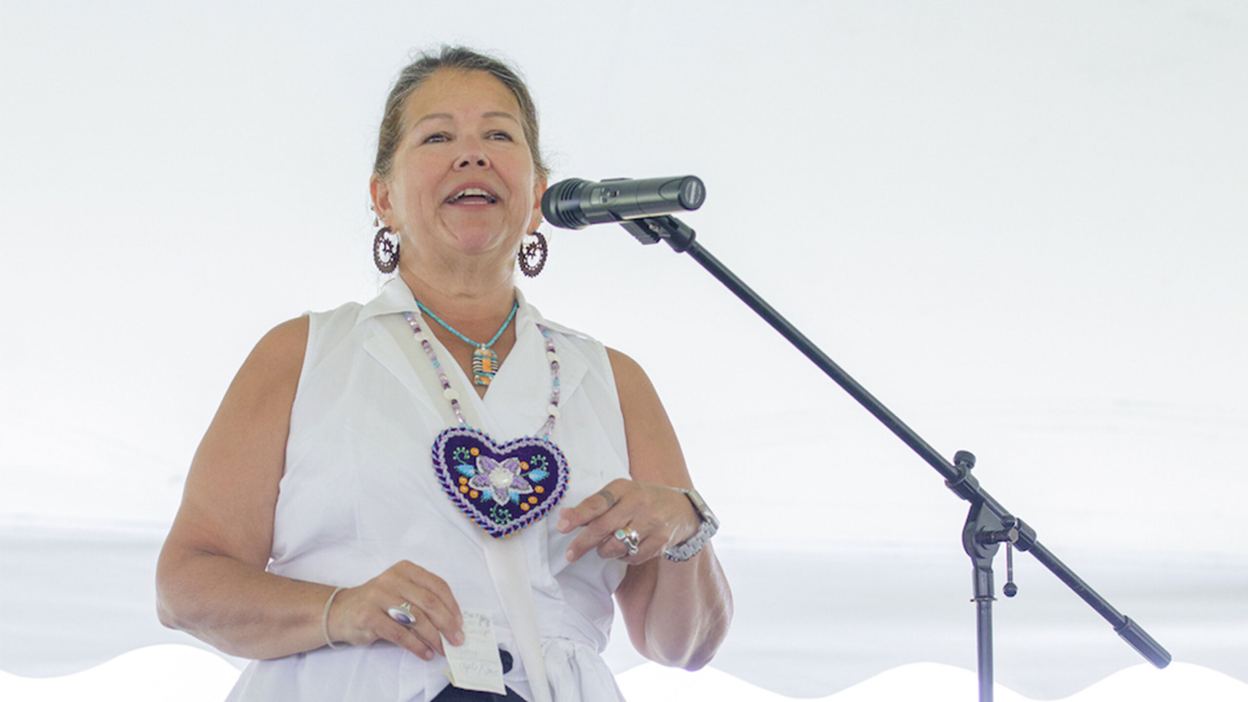 A woman stands on a stage speaking into a microphone, exuding confidence and joy. She wears a sleeveless white top adorned with intricate beadwork around the neckline...