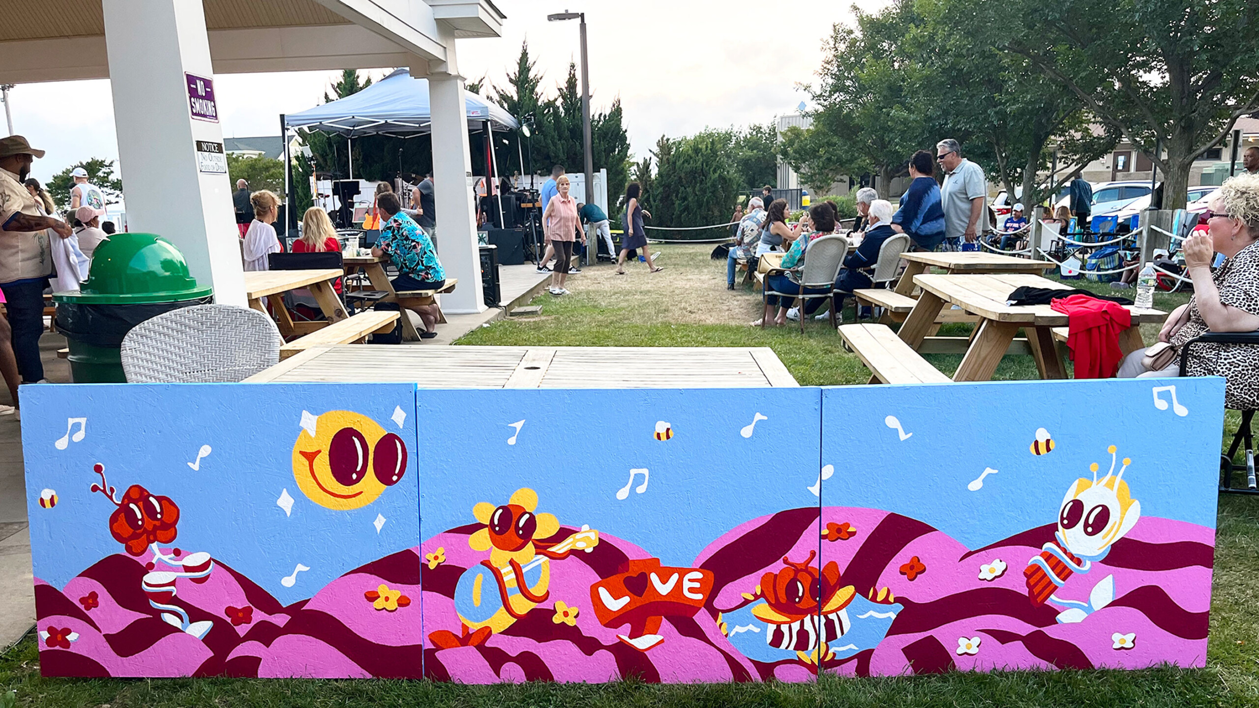 An outdoor festival scene featuring a vibrant, multi-panel art piece in the foreground. The painting is colorful, depicting whimsical cartoon characters resembling...