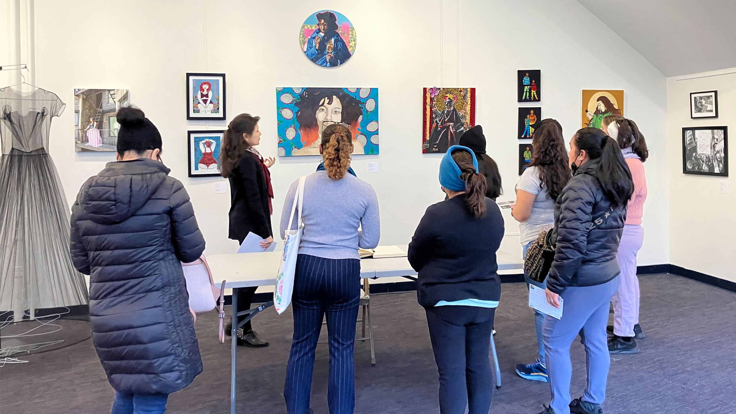A group of eight people is gathered in an art gallery, attentively listening to a guide who is explaining various artworks. The artworks on display include vibrant...