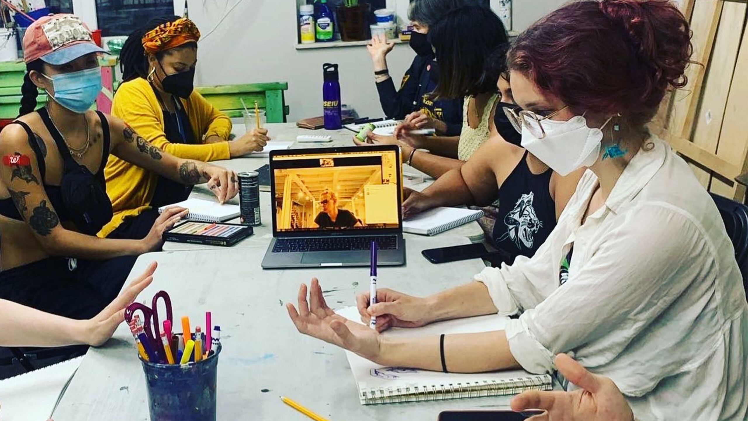 Group of people at a workshop with art supplies and a laptop displaying a presentation.