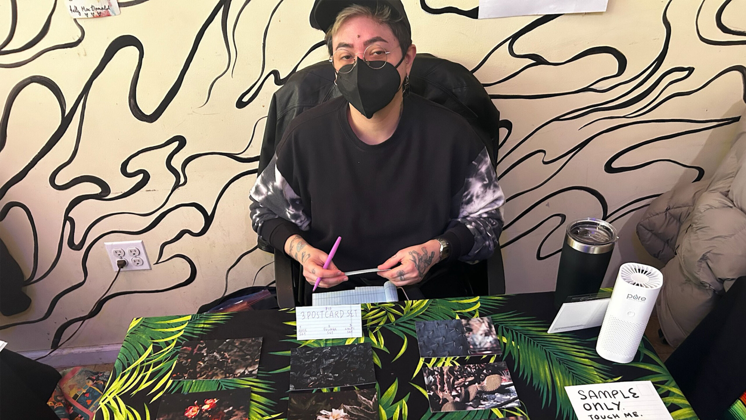 A person is seated behind a table covered with various images of artwork. They are wearing a black face mask and a dark sweater with tie-dye patterns on the sleeves....