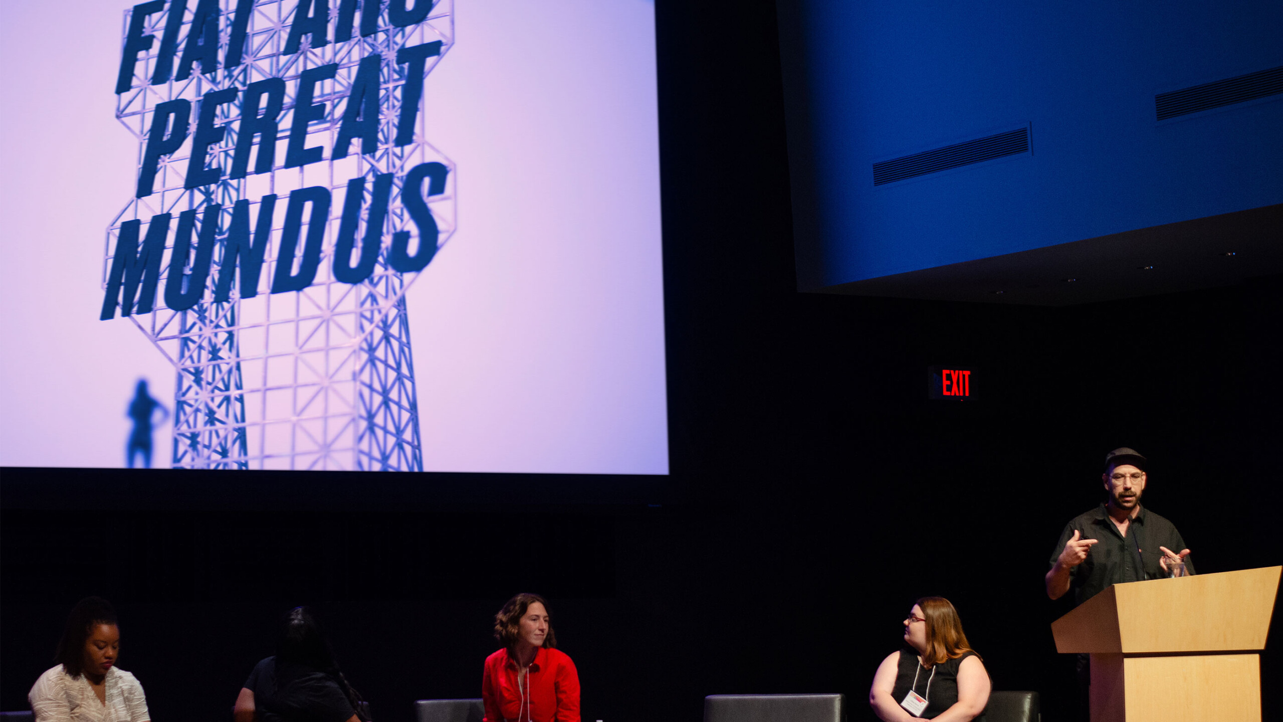 A man is speaking at a wooden podium positioned on the right side of a stage. Four people are seated on chairs, appearing to listen to the speaker. All individuals...