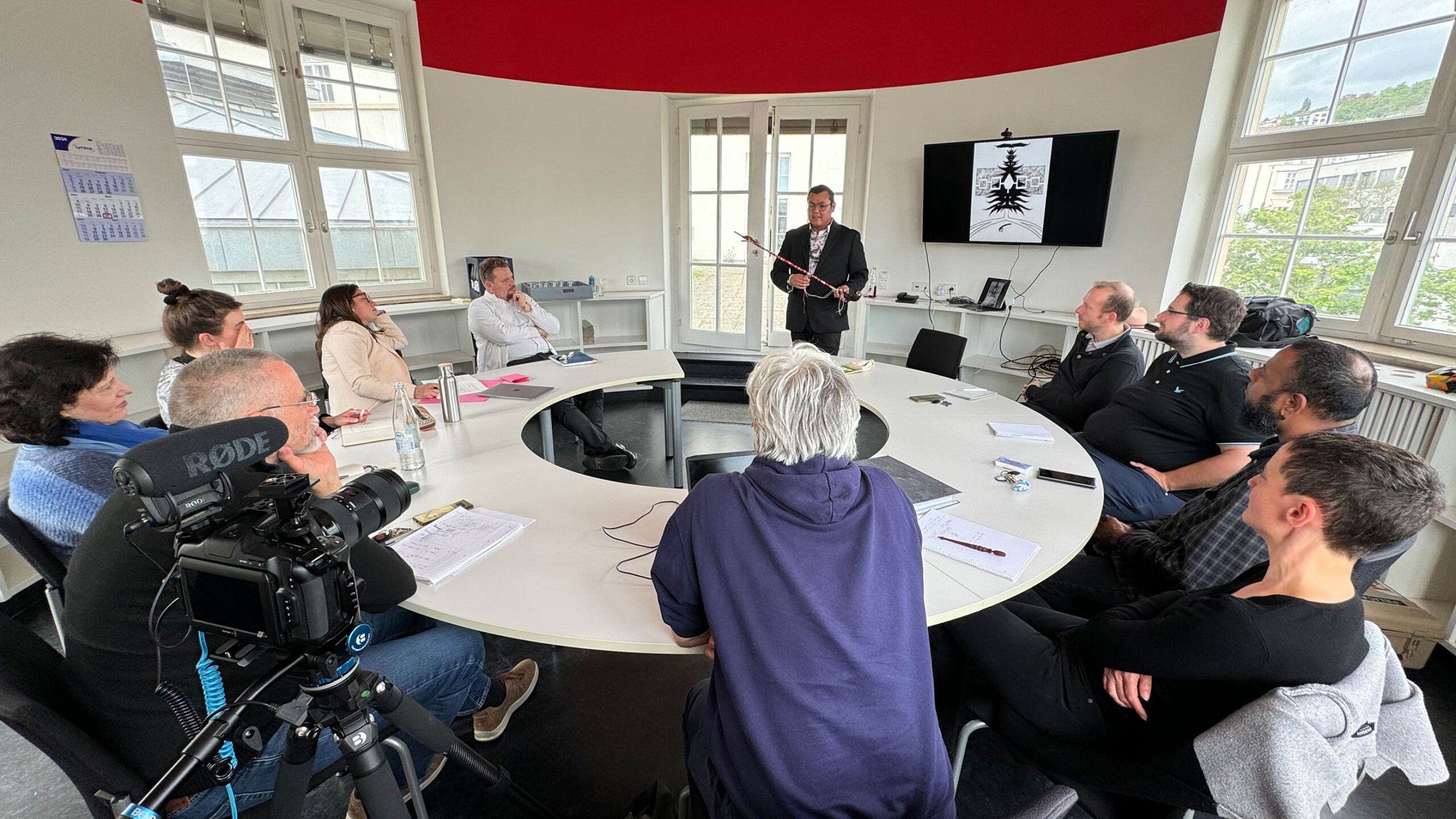 A group of people gathered around a large, oval conference table in a modern meeting room. The room features a distinct red accent at the top of the walls. Most...