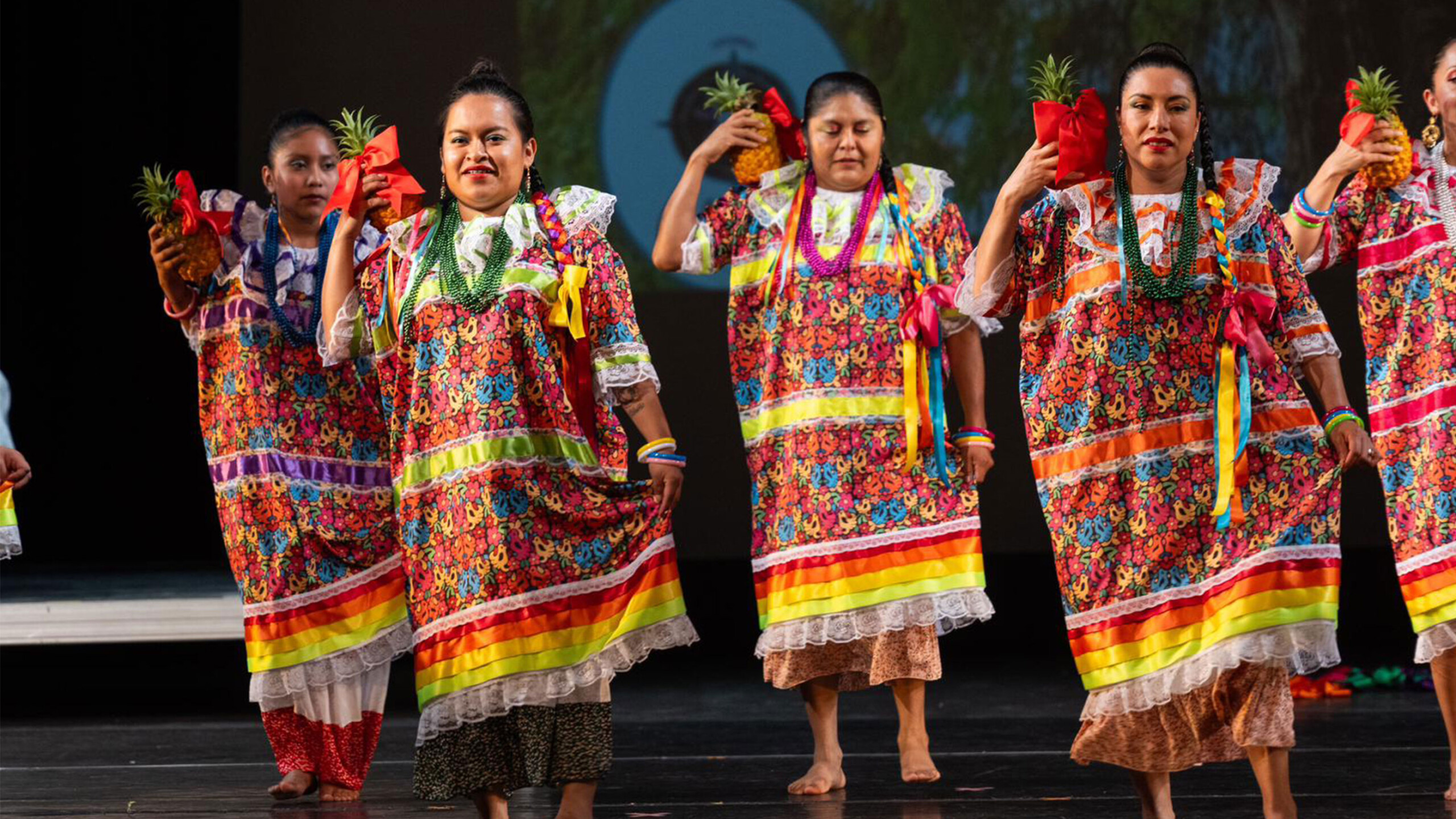 A group of women participates in a cultural dance performance, wearing elaborate traditional Mexican dresses. These vibrant garments feature a multitude of colors...