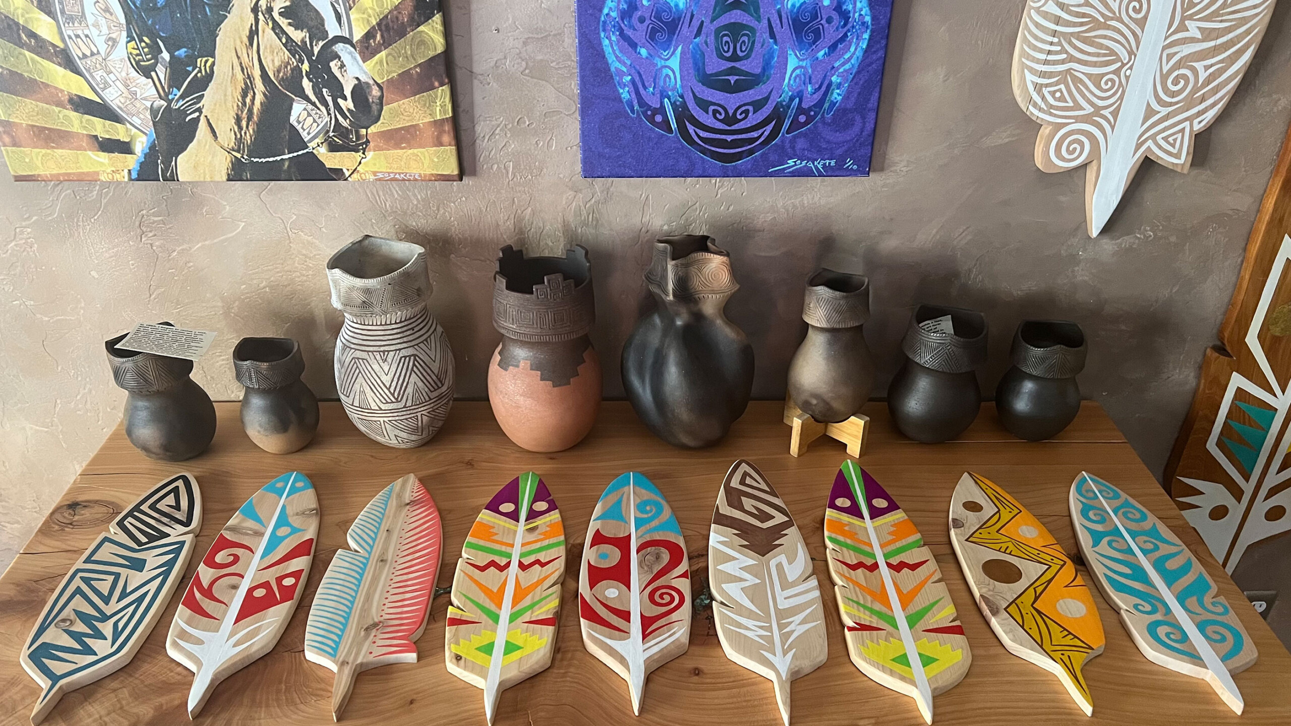 A collection of handmade pottery and intricately painted wooden feathers arranged on a wooden surface. The pottery consists of various vessels in different shapes...