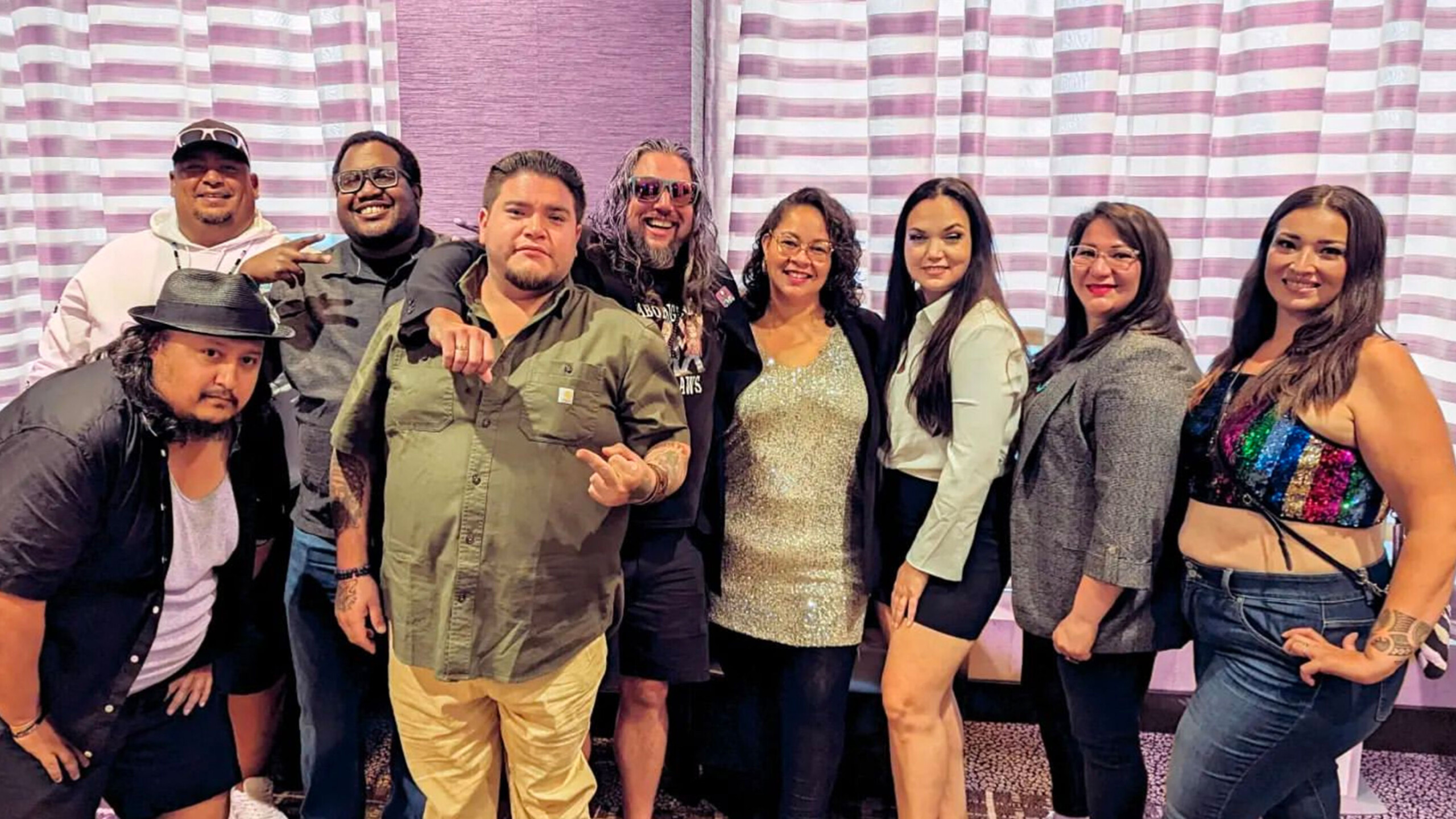 A diverse group of nine individuals stands together indoors, displaying a variety of fashion styles. The background features light-striped curtains and a light...