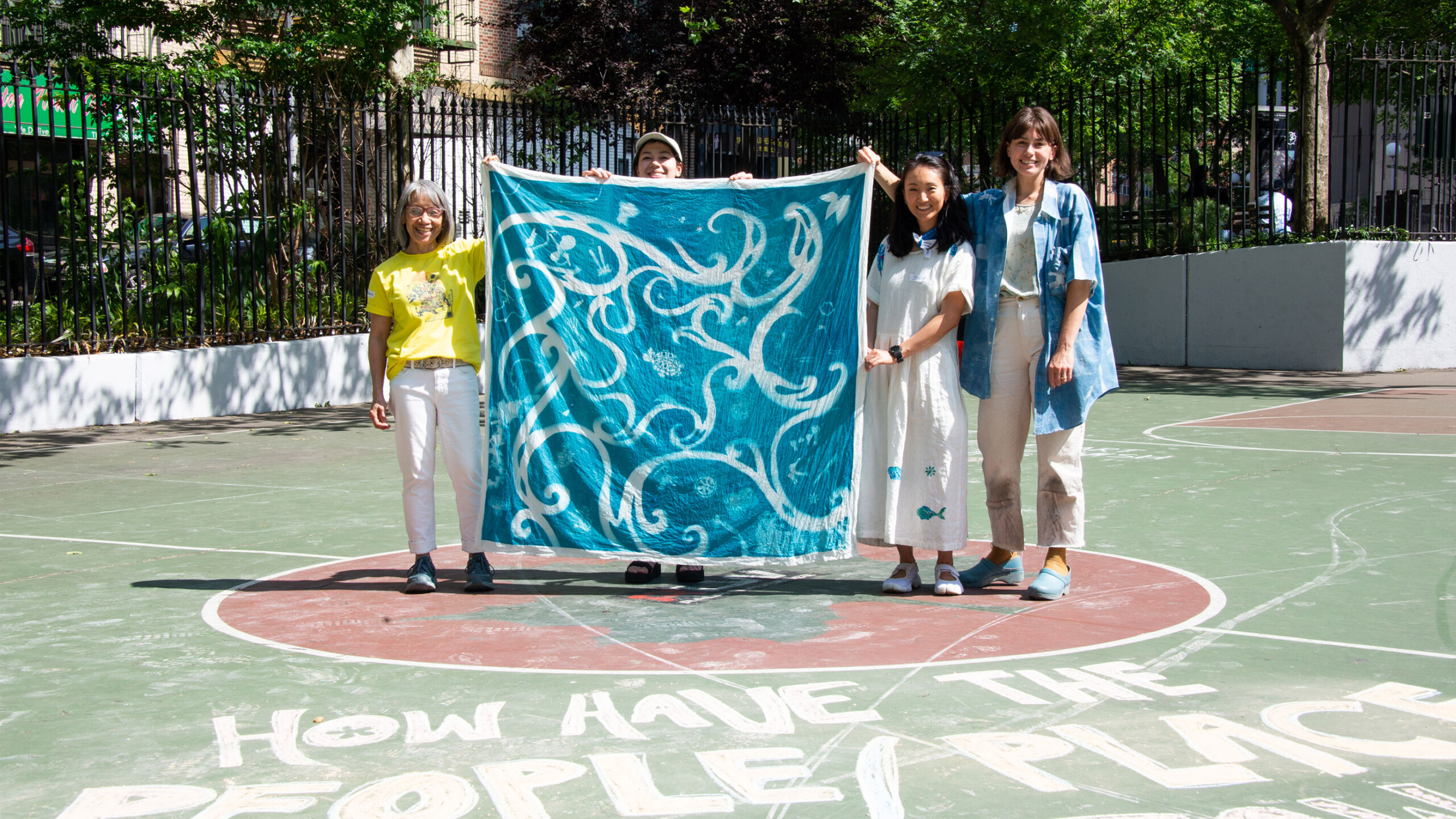 Four people stand on an outdoor basketball court, each holding a corner of a large, blue square cloth adorned with swirling, white patterns. The court's surface...